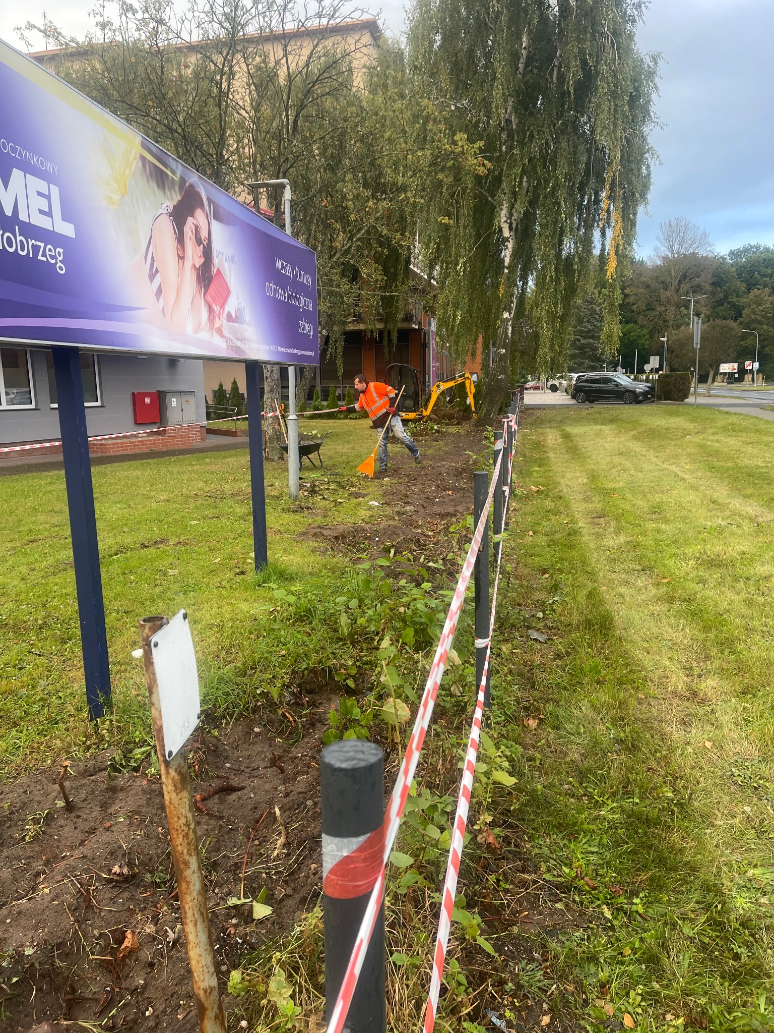 Pracownik w pomarańczowej kamizelce sprząta teren po wycince drzewa przy ulicy, widoczny mały traktor, billboard reklamowy w tle i taśma ostrzegawcza.