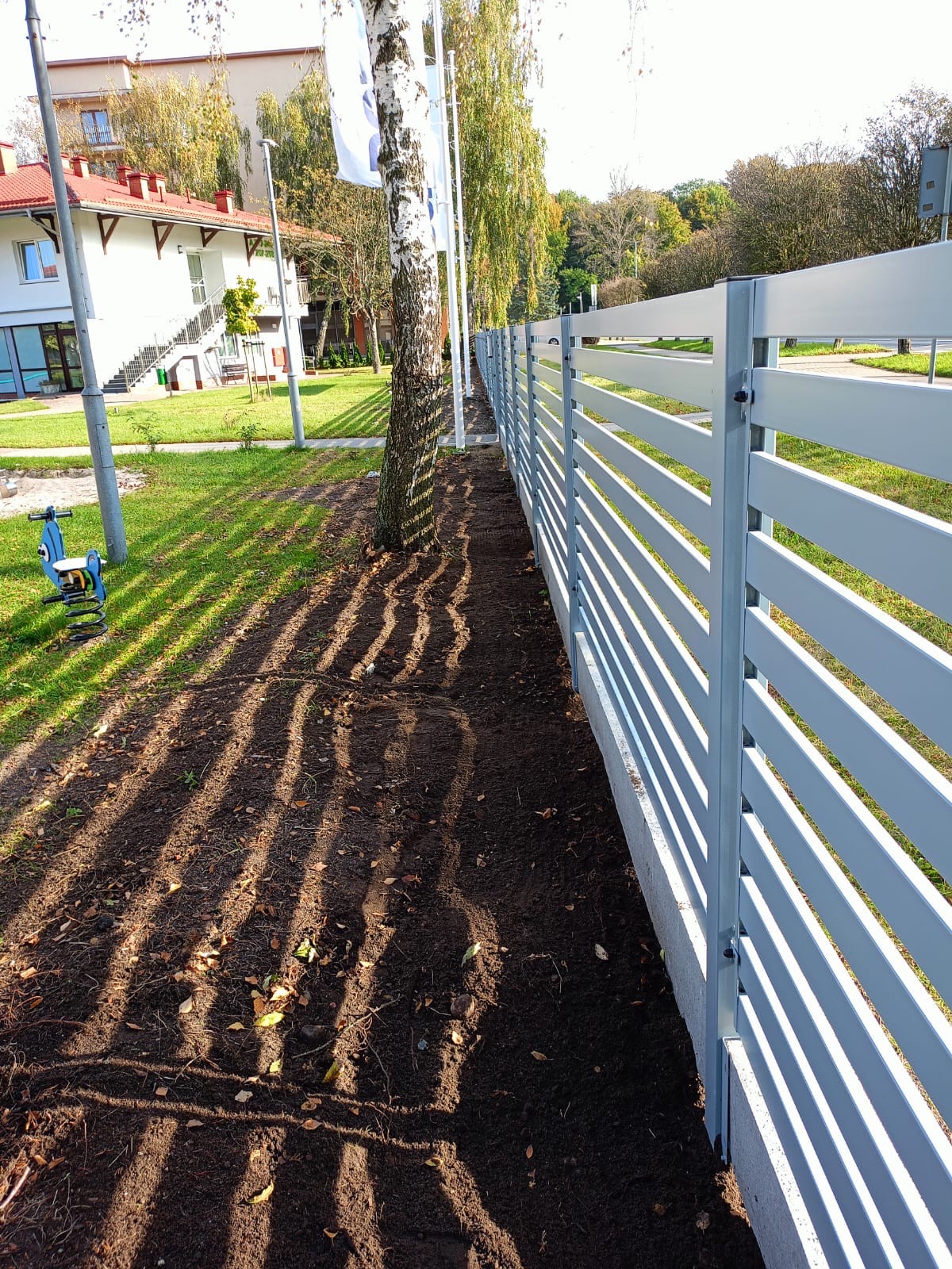 Nowo postawione, jasnoszare, poziome ogrodzenie panelowe z betonu i metalu, oddzielające trawnik od ścieżki, z widocznymi cieniami drzew na ziemi.