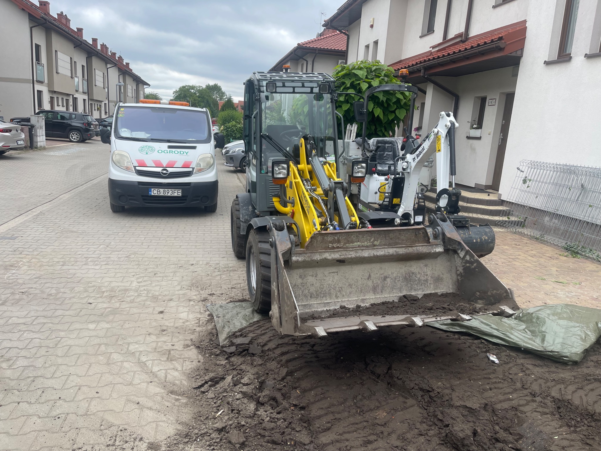 Żółto-szara mini koparka Wacker Neuson na kostce brukowej z wykopaną ziemią, obok biały bus z logo ogrodniczym i budynki mieszkalne w tle.