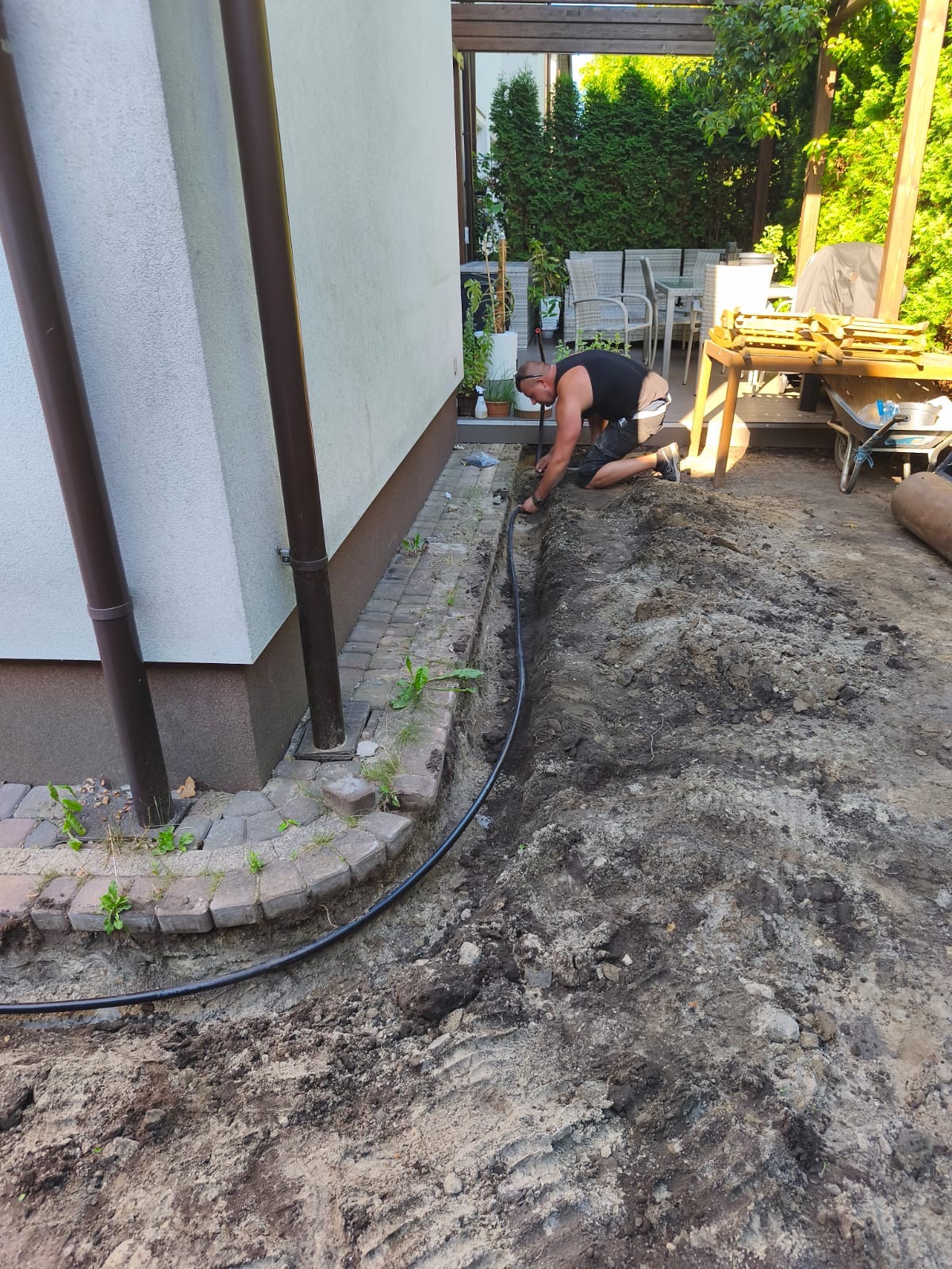 Mężczyzna instaluje system nawadniania ogrodu, układając czarną rurę w wykopanym rowie wzdłuż ściany domu z rynnami. Widoczne narzędzia i częściowo wyposażony taras.