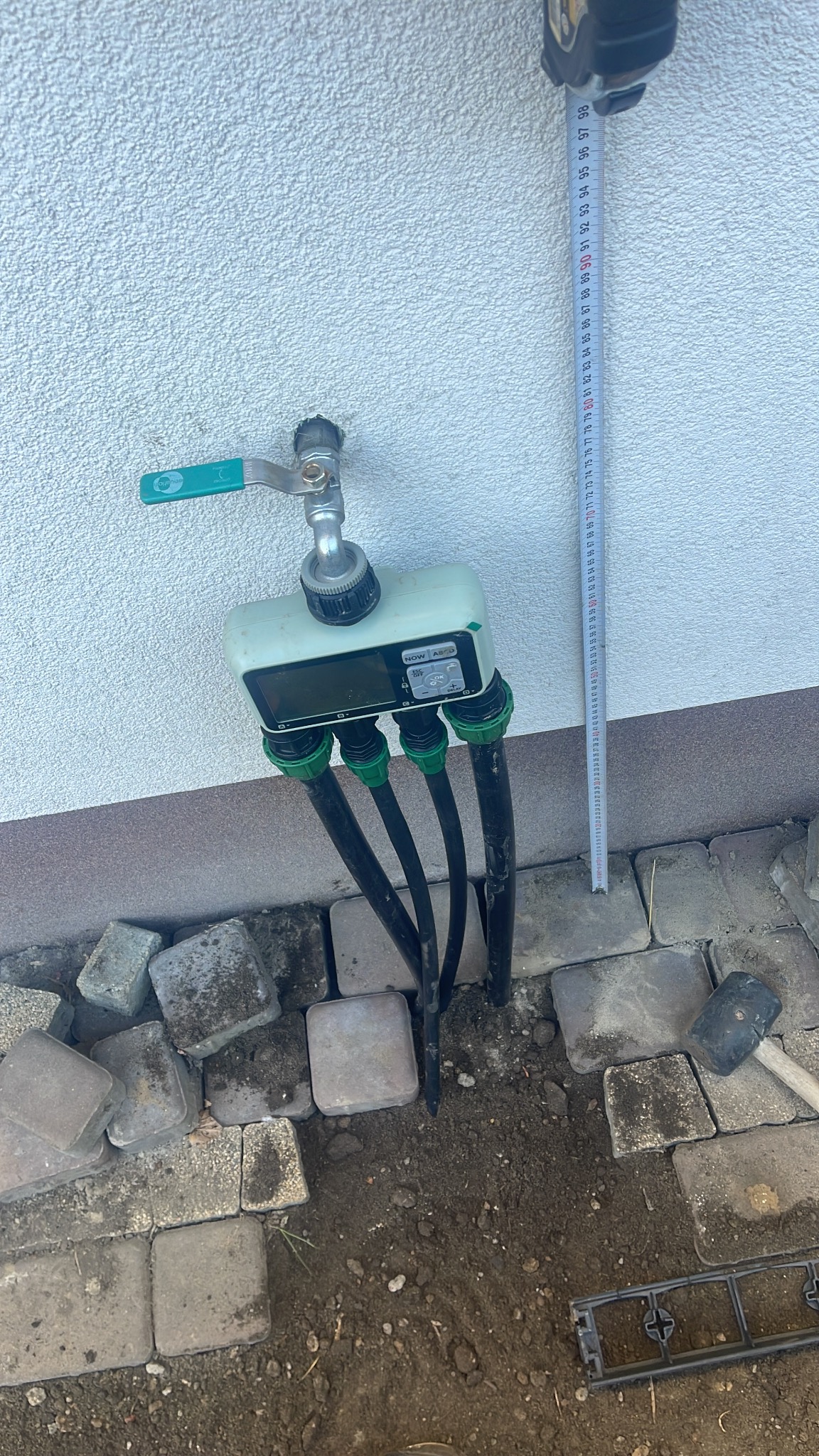 Podłączenie systemu nawadniania: kran z elektronicznym sterownikiem i czterema wężami, obok leżą kostki brukowe i miara zwinięta na ścianie.
