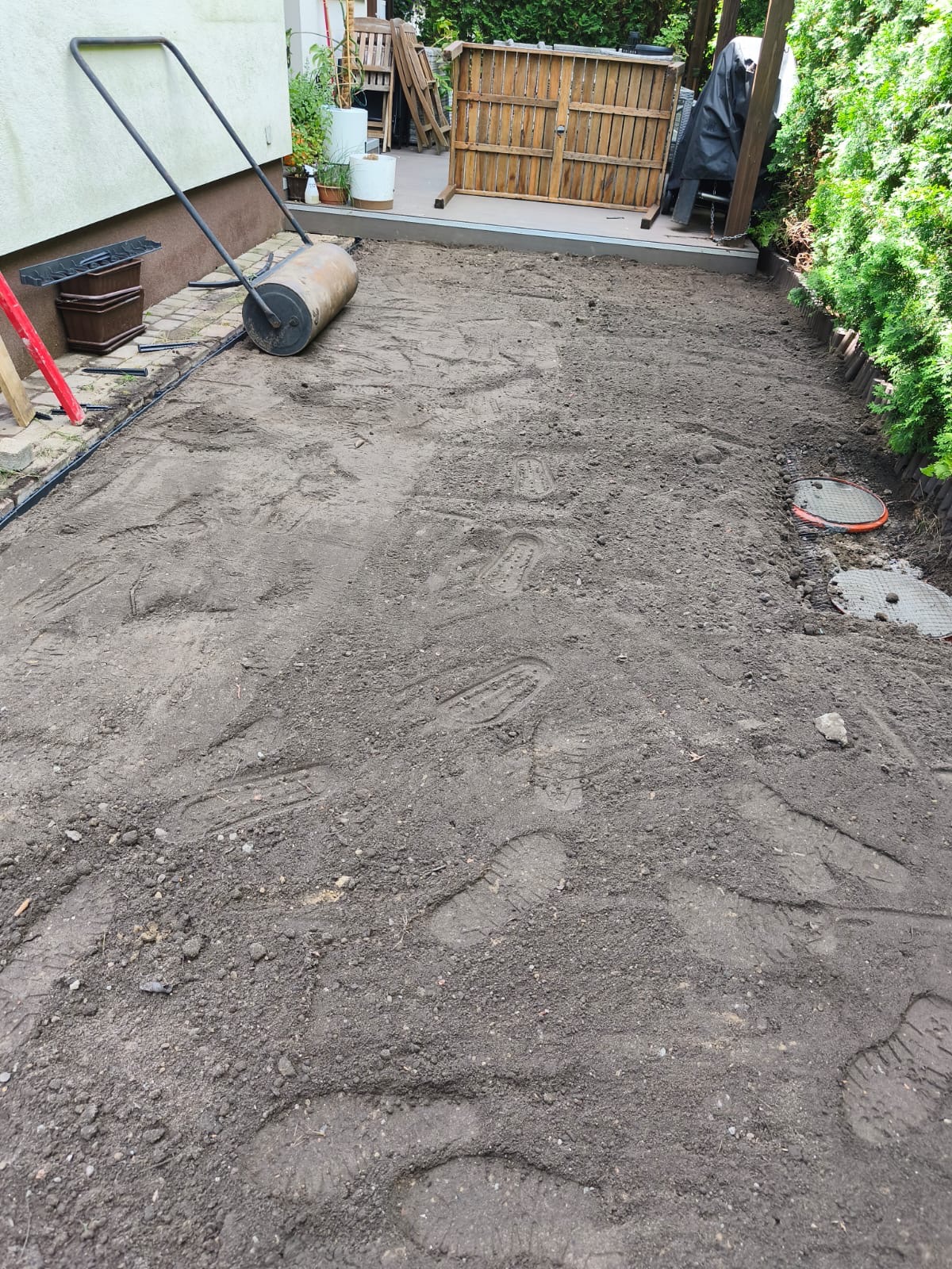 Przygotowany teren pod trawnik z widocznymi śladami stóp i walcem ogrodowym. W tle drewniana konstrukcja i zieleń. Etap wyrównywania gruntu.