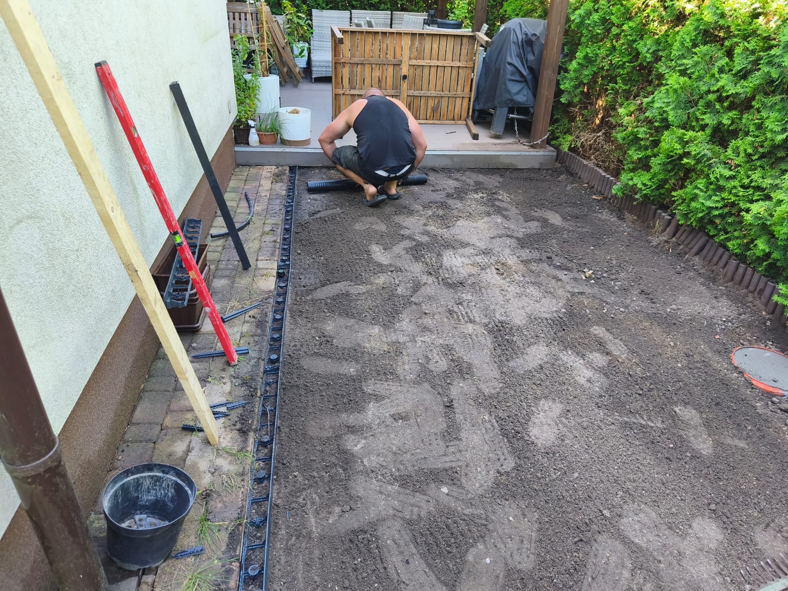 Prace przygotowawcze pod trawnik: mężczyzna instaluje obrzeża trawnikowe na wyrównanym terenie. Po lewej narzędzia i poziomice, w tle roślinność i elementy wyposażenia ogrodu.