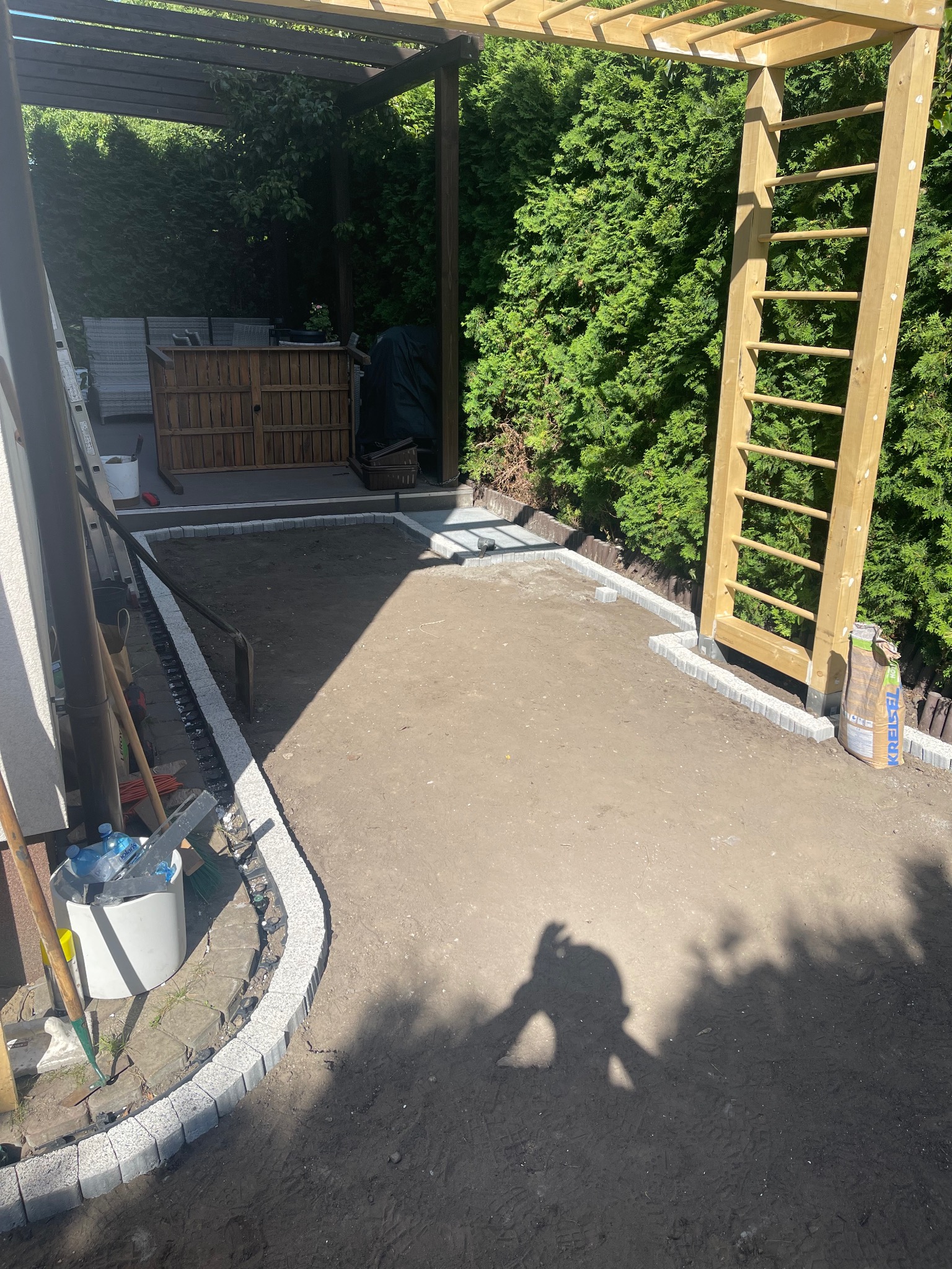 Przygotowanie podłoża pod trawnik z obrzeżami z kostki, widoczne narzędzia i cień osoby. Etap prac w ogrodzie przed założeniem trawy.