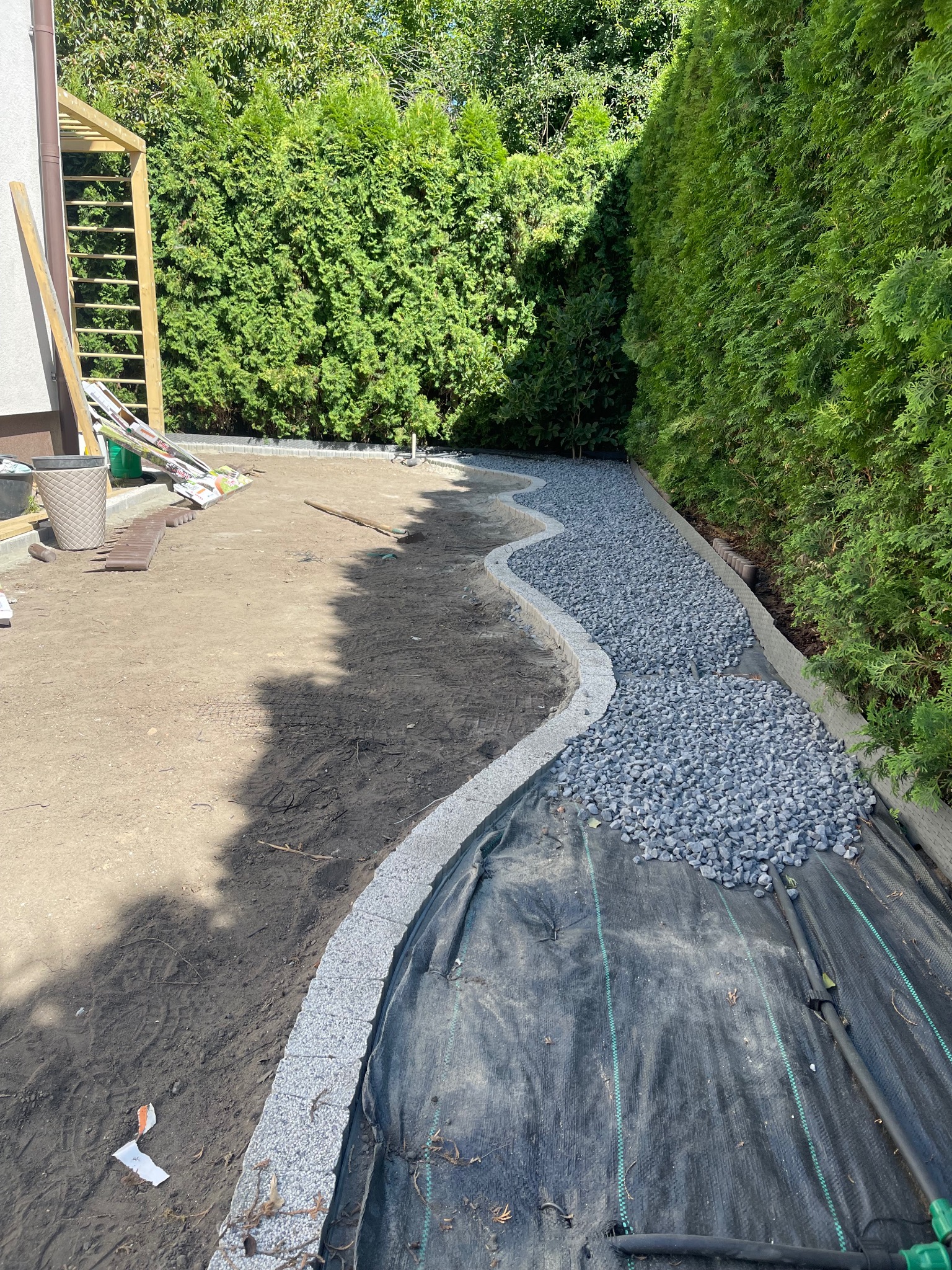 Kształtowanie ścieżki w ogrodzie: obrzeża z kostki, czarna agrowłóknina i wysypany żwir, w tle zielony żywopłot. Etap prac ogrodowych w słoneczny dzień.