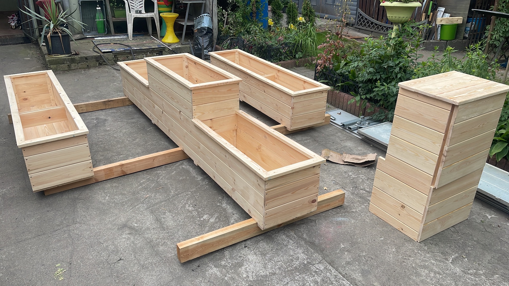 Ręcznie robione, surowe drewniane skrzynie na rośliny ogrodowe, ustawione na betonowym podłożu, z widocznymi elementami konstrukcyjnymi i zielenią w tle.