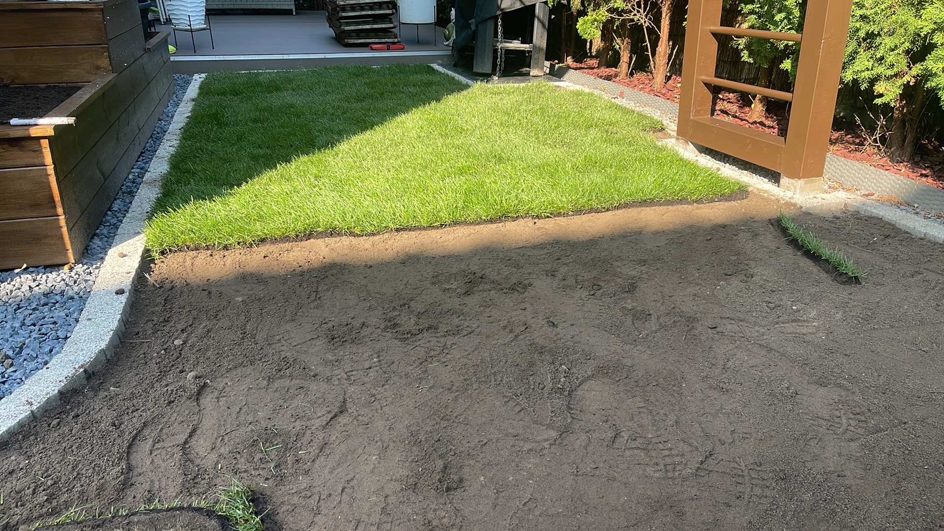 Fragment układania trawy z rolki w ogrodzie. Częściowo ułożona, intensywnie zielona trawa sąsiaduje z przygotowaną glebą. Widoczne obrzeża i elementy architektury ogrodowej.