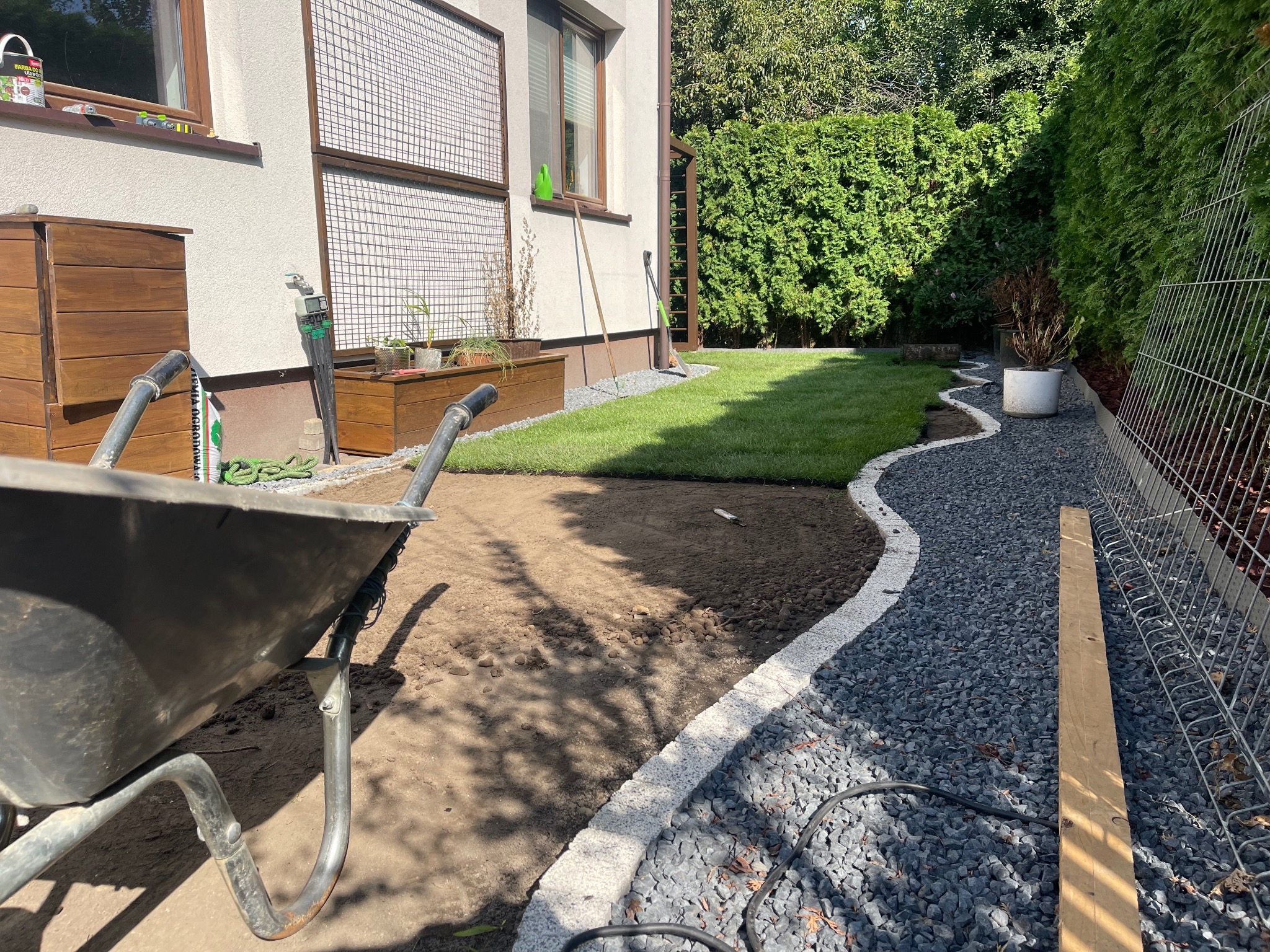 Fragment ogrodu w trakcie realizacji: ułożona trawa, wytyczona ścieżka z kamienia, widoczna taczka i narzędzia ogrodnicze. Częściowo przygotowany teren pod dalsze prace.