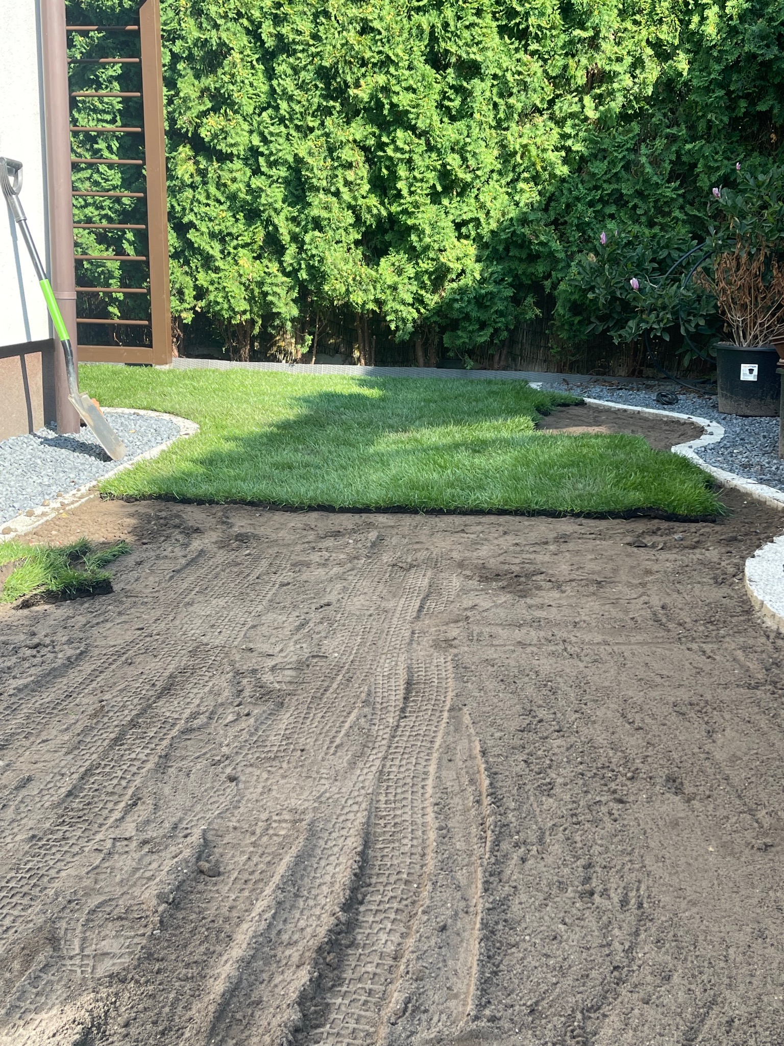 Układanie trawy z rolki w ogrodzie. Widoczne fragmenty ułożonej trawy, przygotowane podłoże z wyraźnymi śladami opon i narzędzia ogrodnicze.