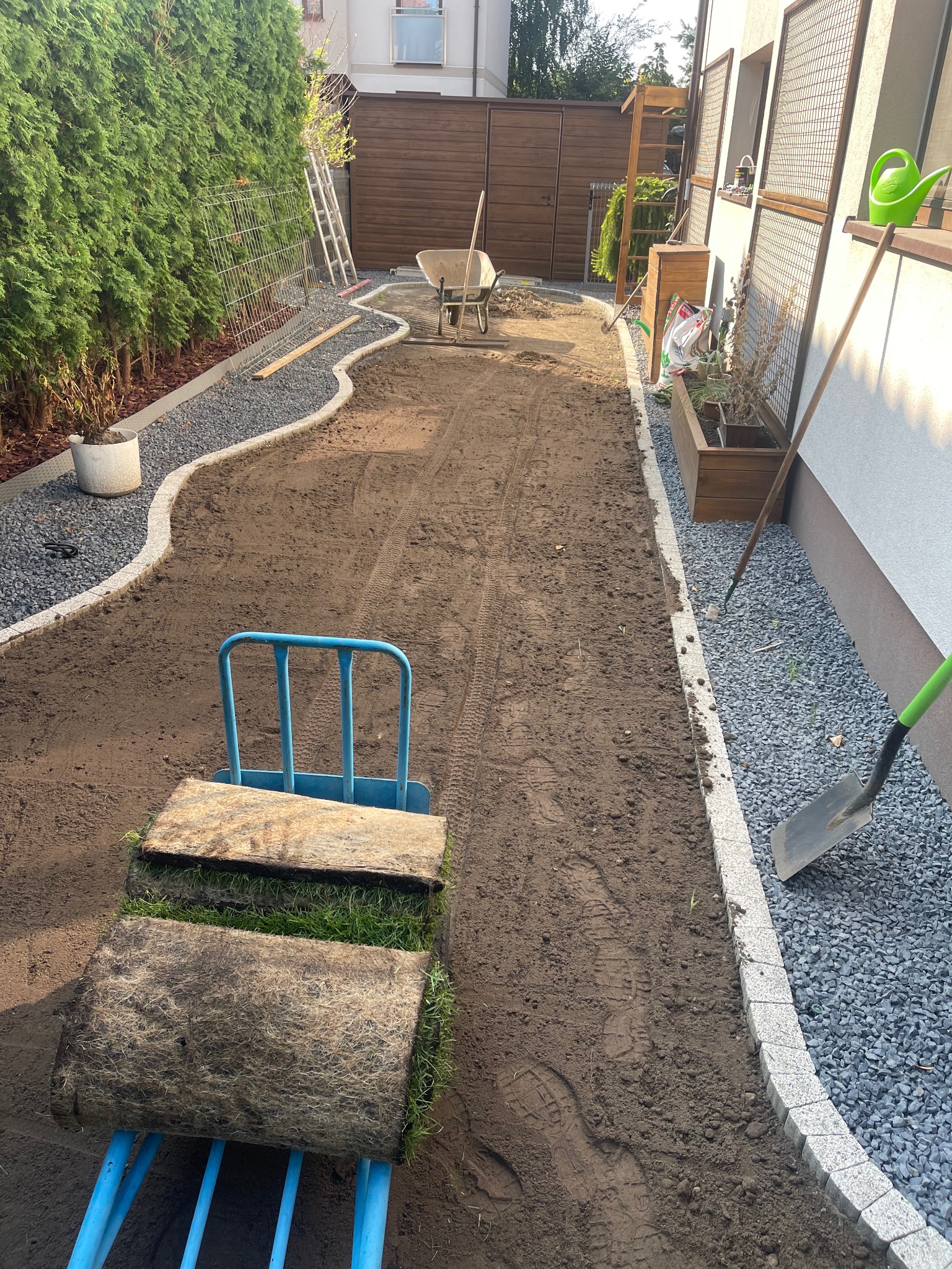 Przygotowany teren pod trawnik z rolki, widoczny wałek z trawą na wózku, obrzeża z kamieni i narzędzia ogrodnicze. Etap prac w ogrodzie.