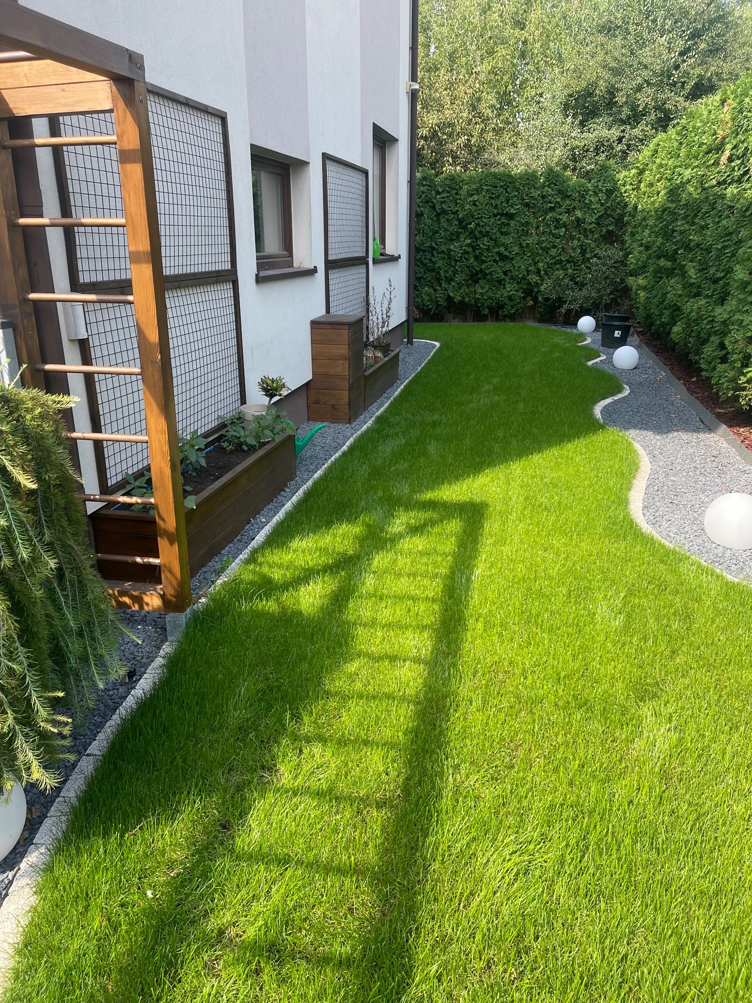 Zadbany trawnik z rolki, obrzeża z kamienia, drewniane donice, pergola i kuliste lampy ogrodowe w słonecznym ogrodzie przy białym domu.