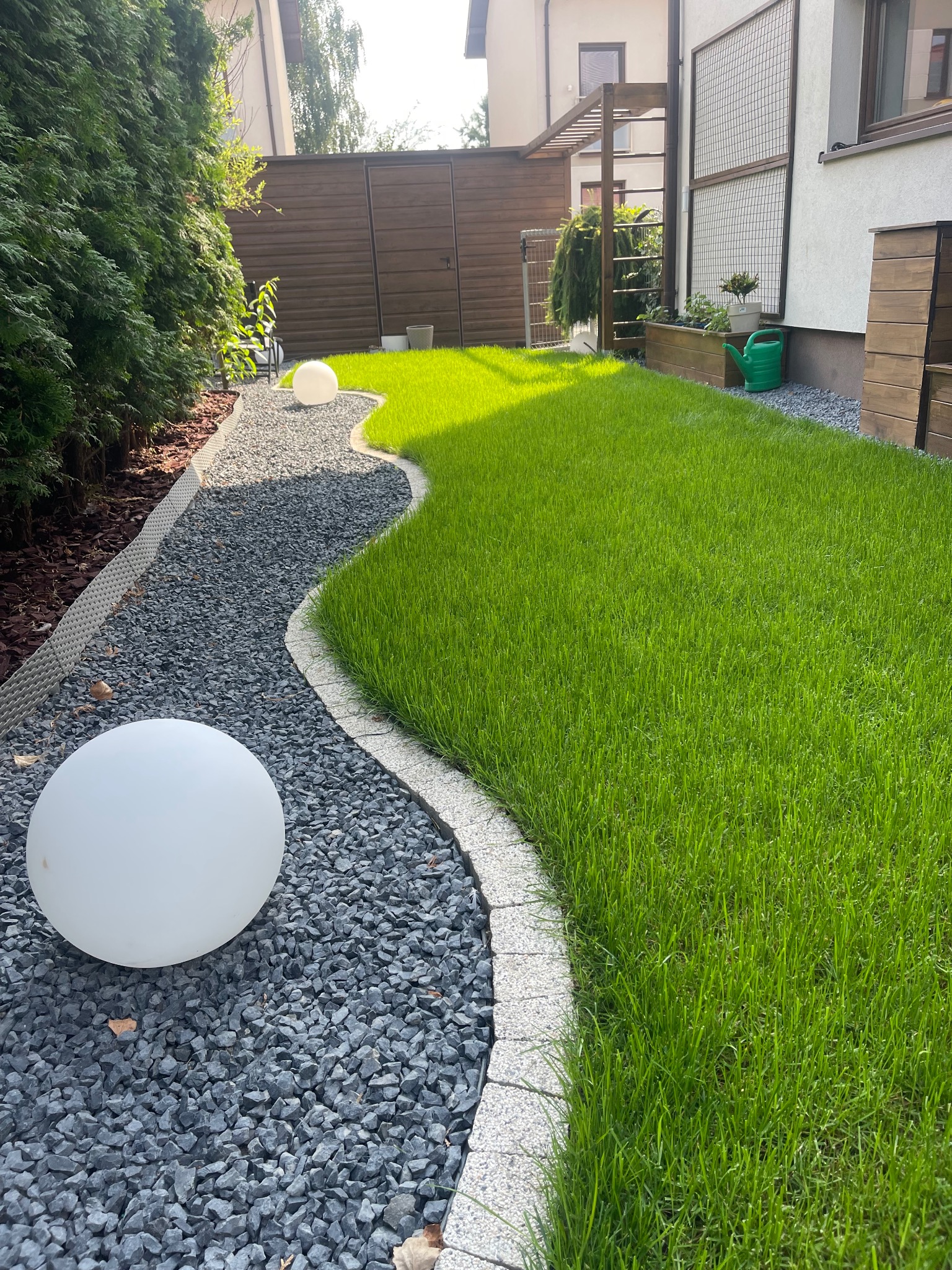 Falista ścieżka z szarego żwiru, obrzeża z kostki, trawnik z soczystą zielenią, oświetlony kulami, w tle drewniane ogrodzenie i pergola.