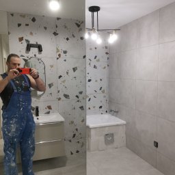 Remontsado - Mężczyzna w ubraniu roboczym robi zdjęcie telefonem w nowo wykończonej łazience z umywalką i wanną wyłożoną szarymi płytkami. Po lewej stronie ściana wykończona płytkami w mozaikę.