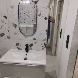 Remontsado - Nowoczesna umywalka z czarną armaturą na tle ściany wyłożonej płytkami imitującymi lastryko, lustro w czarnej ramie, widoczne prace wykończeniowe przy ościeżnicy drzwiowej.