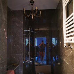 Remontsado - Wykończona łazienka z ciemnymi płytkami imitującymi marmur, widoczna bateria prysznicowa, grzejnik drabinkowy, ozdobny żyrandol i odbicie osoby robiącej zdjęcie. W tle widoczne kartony i narzędzia.