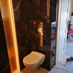 Nowoczesna łazienka z czarnymi marmurowymi płytkami, podświetlanym panelem dekoracyjnym o strukturze plastra miodu i podwieszaną toaletą.