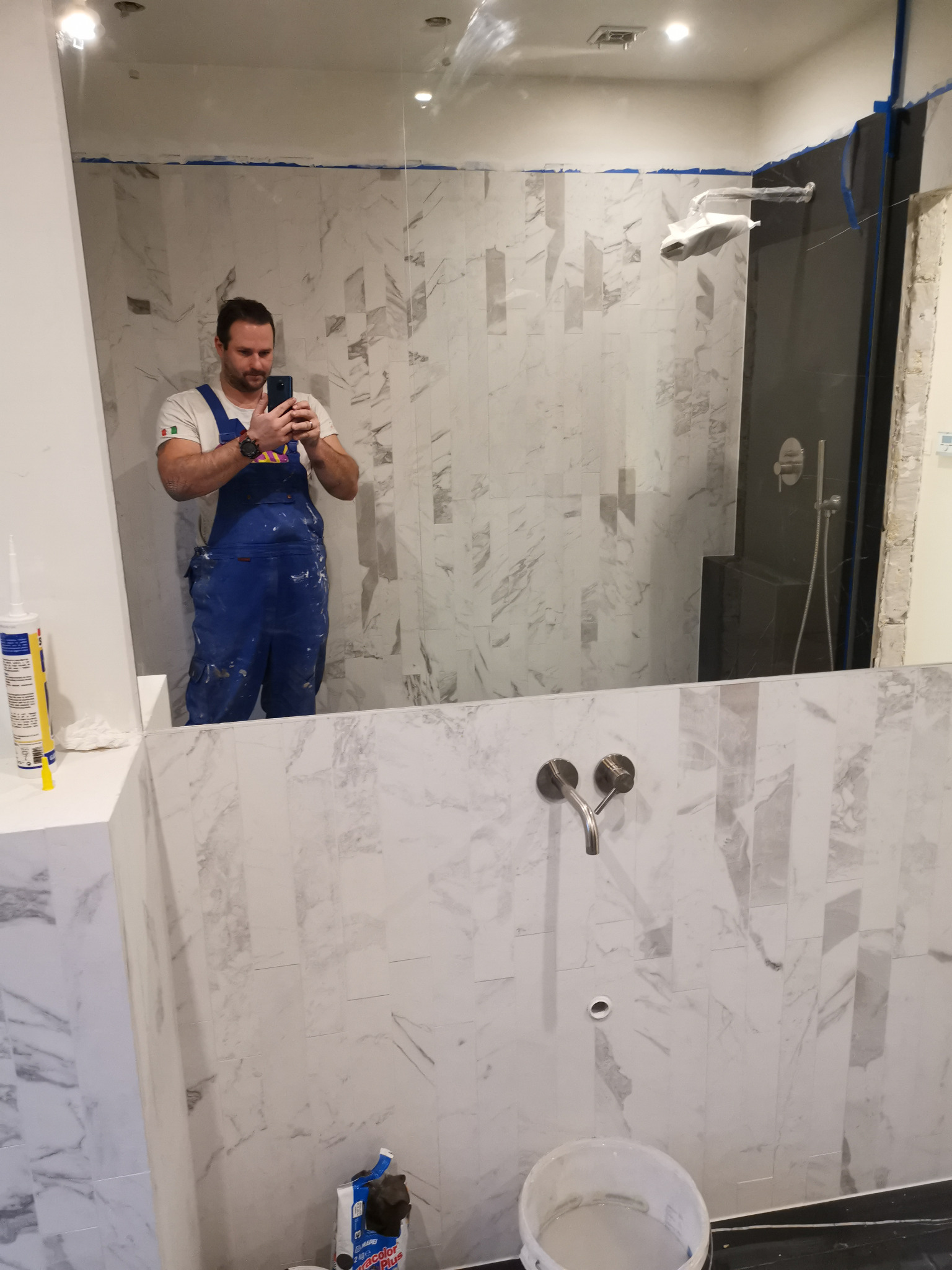 Autoportret glazurnika w lustrze podczas remontu łazienki, widoczne płytki imitujące marmur, bateria umywalkowa, kabina prysznicowa w folii ochronnej oraz narzędzia i materiały budowlane.