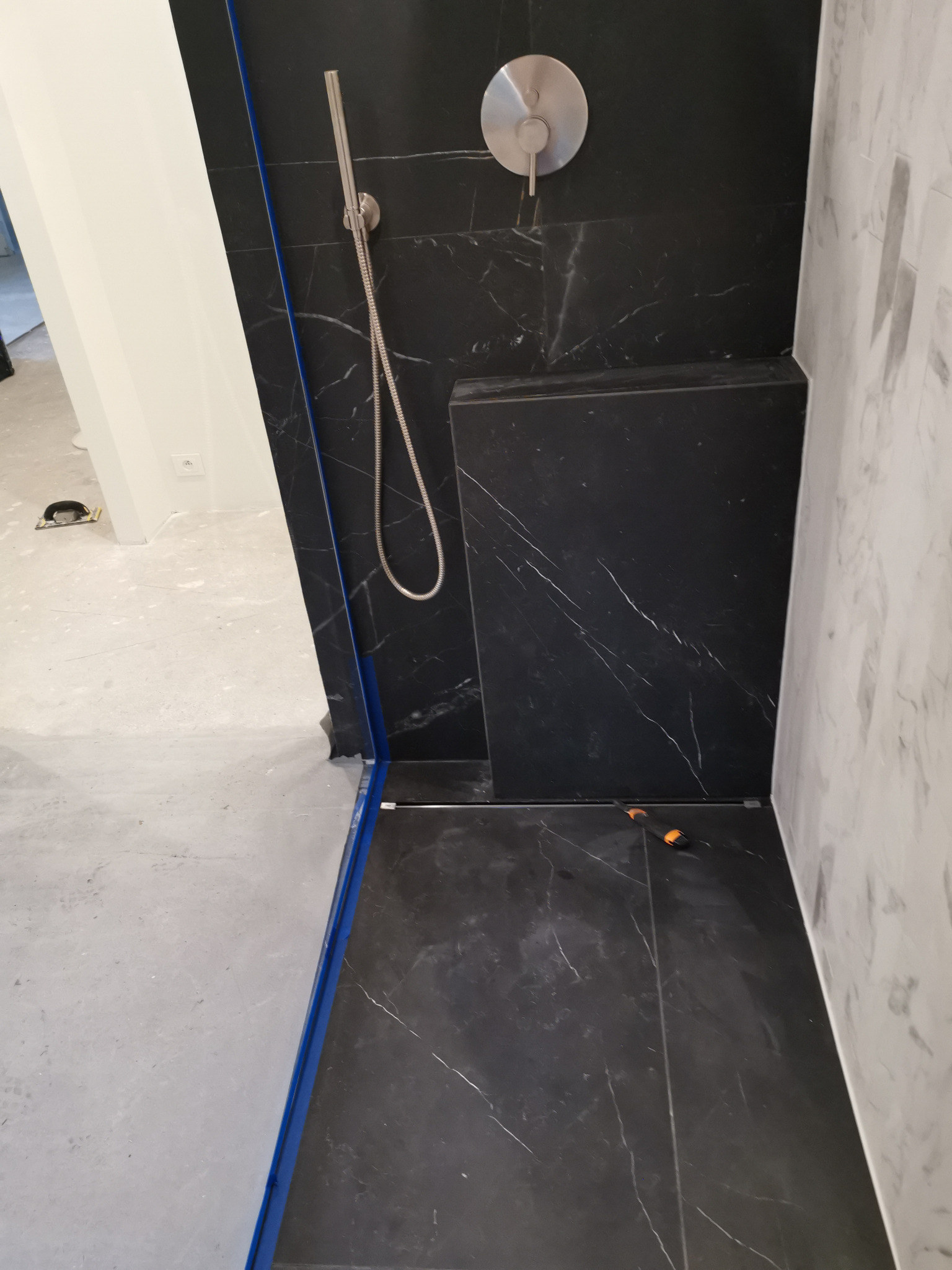 Niedokończona kabina prysznicowa z czarnymi płytkami imitującymi marmur, widoczny odpływ liniowy, bateria prysznicowa i białe płytki na sąsiedniej ścianie, podłoga surowa, narzędzie glazurnicze...