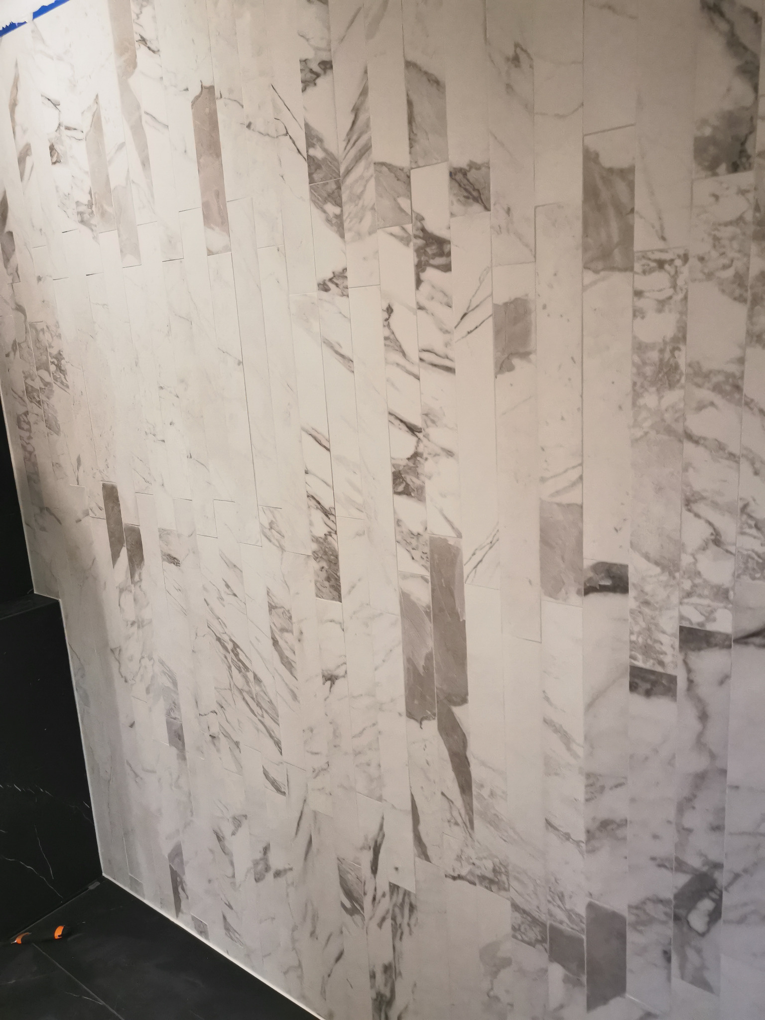Ściana wyłożona wąskimi, pionowymi płytkami imitującymi biały marmur z szarymi żyłkami, przylegająca do czarnej, marmurowej podłogi, widoczne pomarańczowe kliny dystansowe.