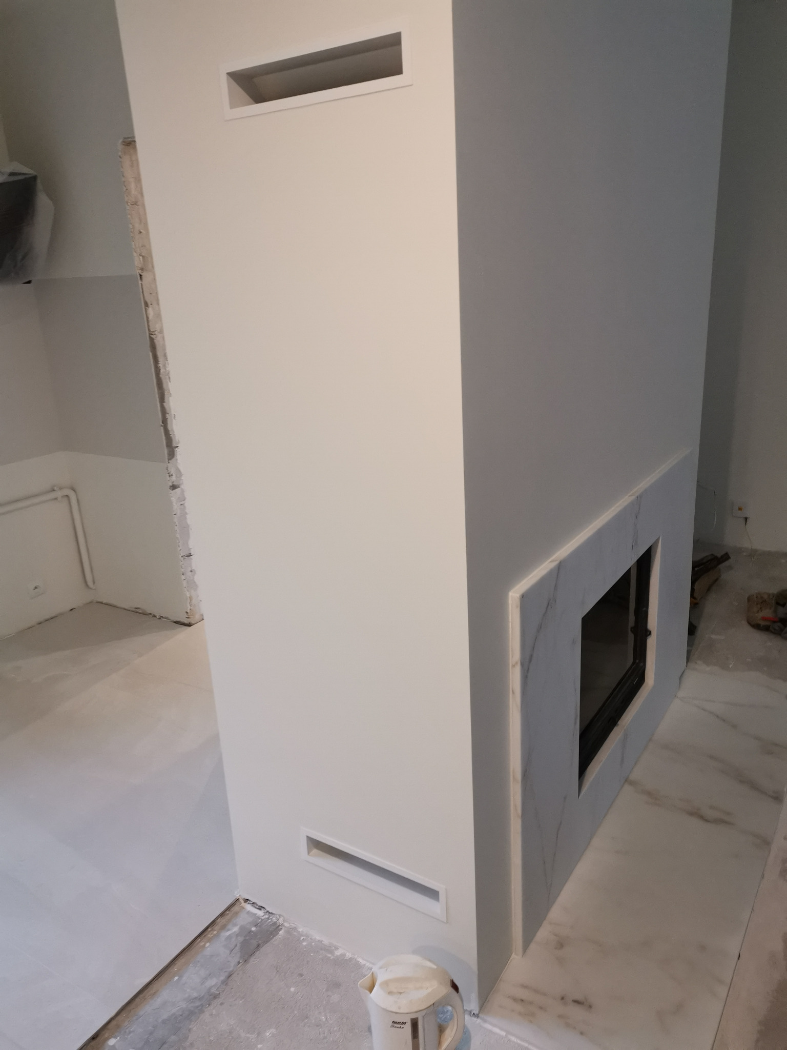 Wykończony kominek z marmurową obudową w jasnym pomieszczeniu, widoczne kratki wentylacyjne w ścianie i fragment podłogi w trakcie prac wykończeniowych.