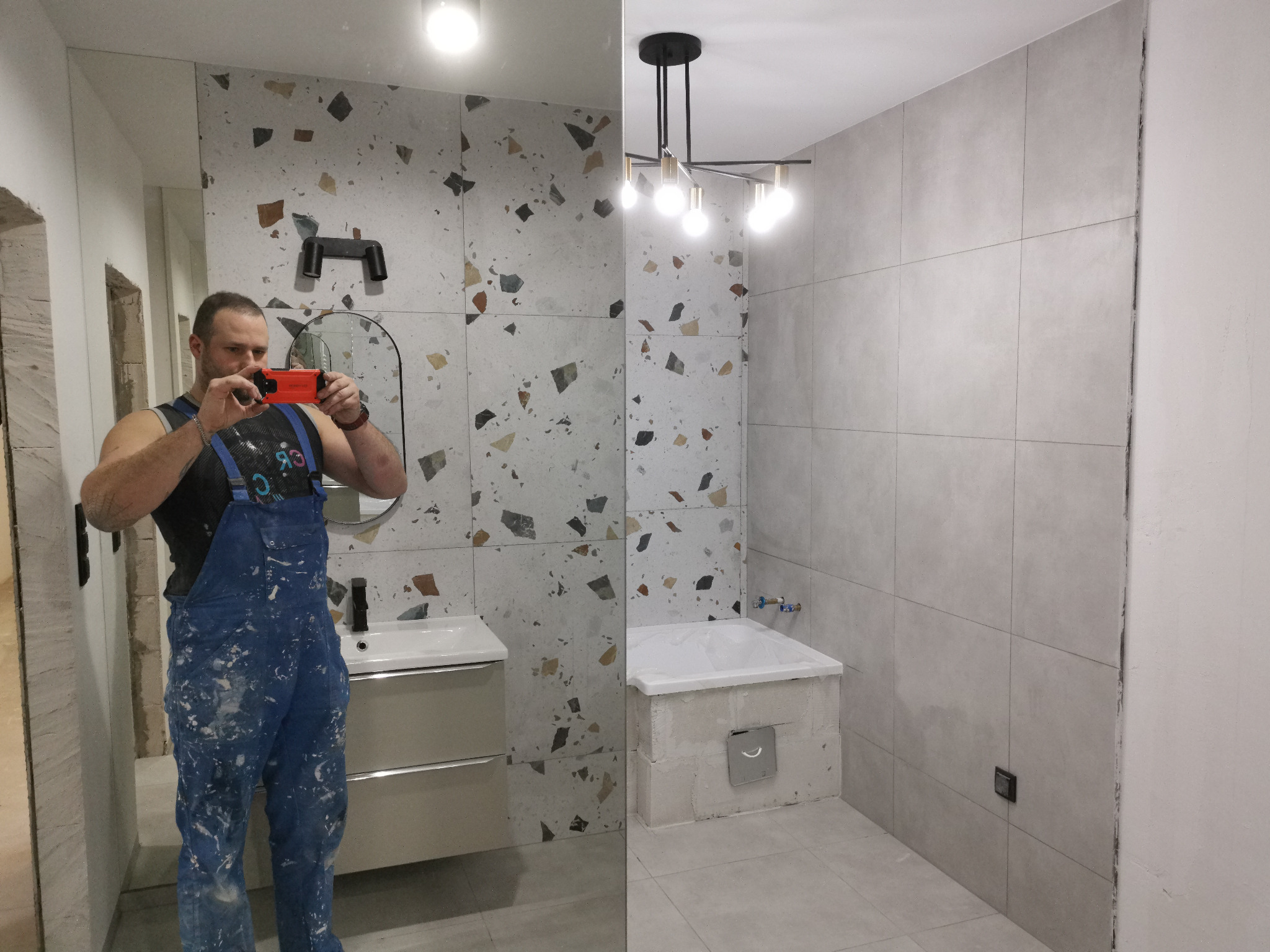 Mężczyzna w ubraniu roboczym robi zdjęcie telefonem w nowo wykończonej łazience z umywalką i wanną wyłożoną szarymi płytkami. Po lewej stronie ściana wykończona płytkami w mozaikę.
