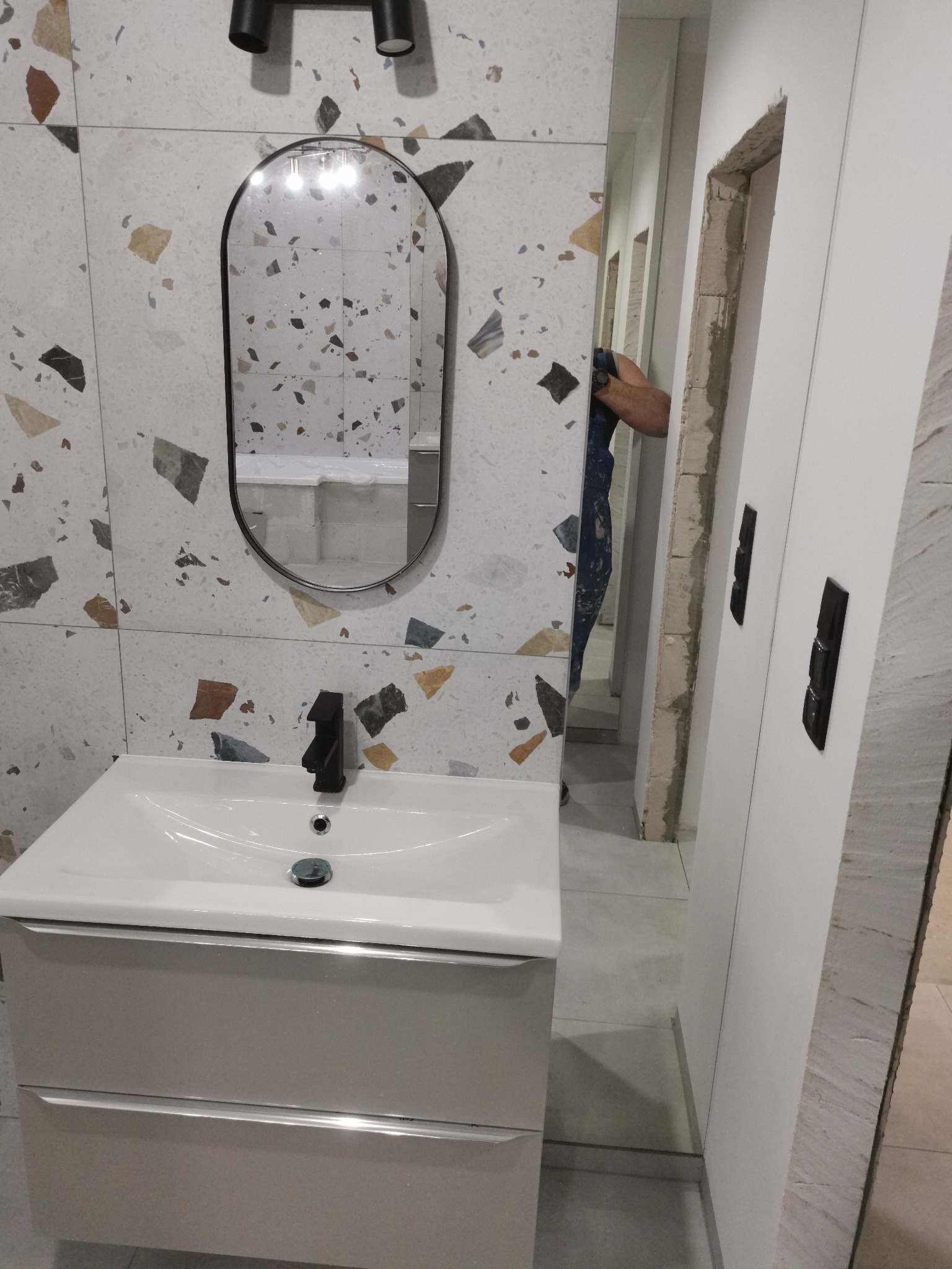 Nowoczesna umywalka z czarną armaturą na tle ściany wyłożonej płytkami imitującymi lastryko, lustro w czarnej ramie, widoczne prace wykończeniowe przy ościeżnicy drzwiowej.