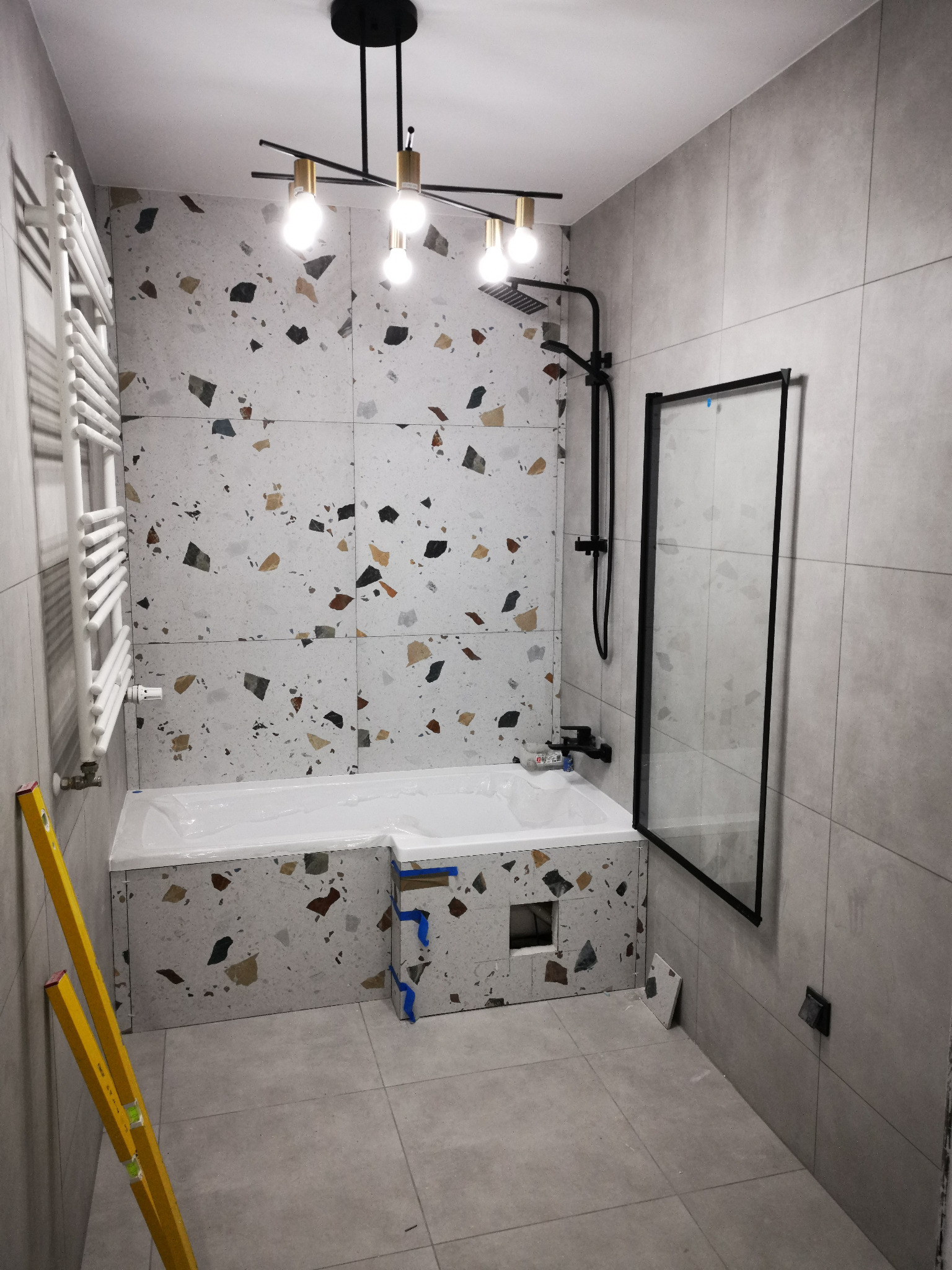 Nowoczesna łazienka w trakcie wykańczania, z wanną obudowaną płytkami imitującymi lastryko, czarną armaturą i szklanym panelem prysznicowym, widoczne poziomice przy ścianie.