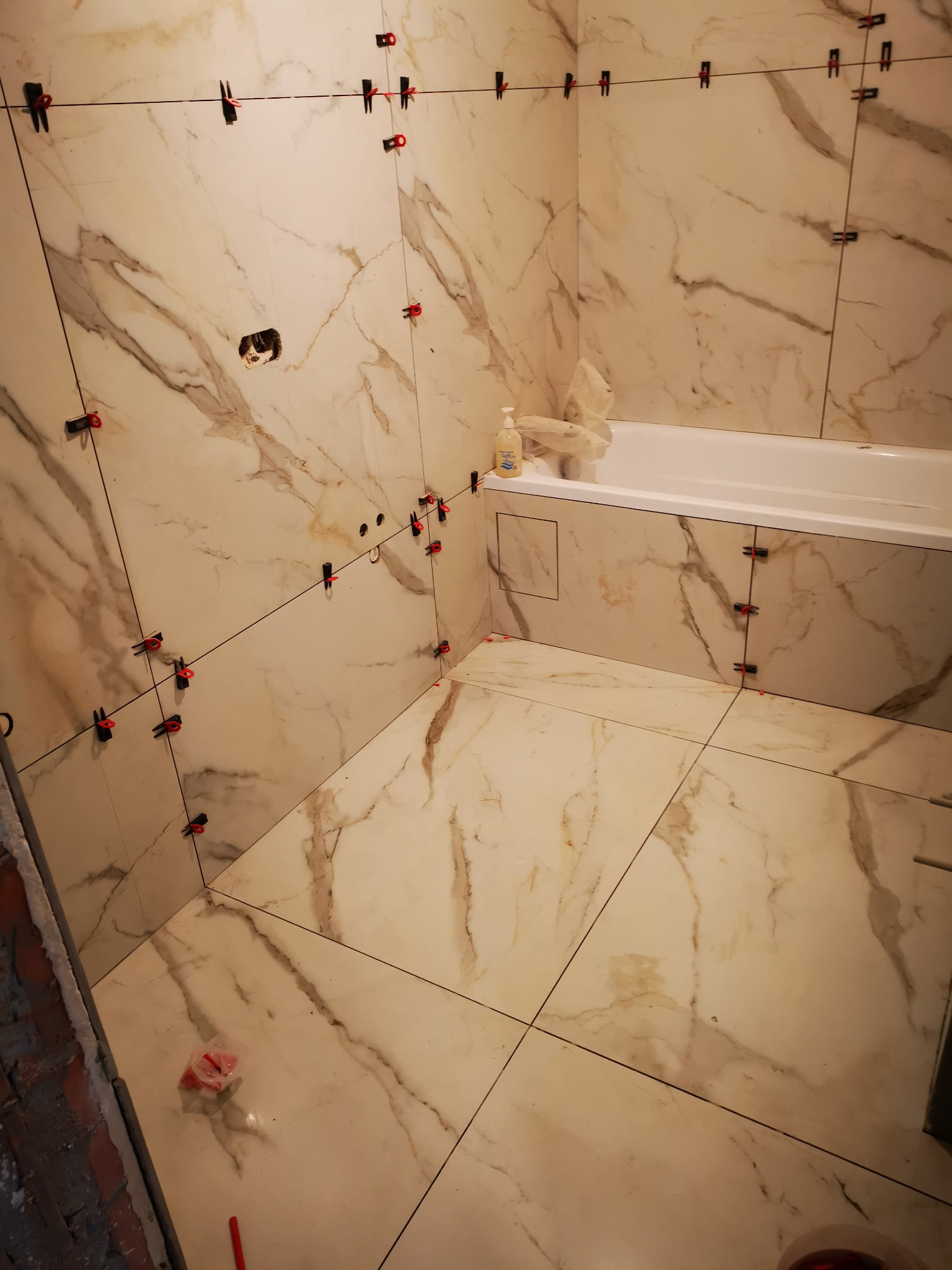 Łazienka w trakcie układania płytek imitujących marmur, widoczne klipsy poziomujące na ścianach i podłodze, fragment wanny i butelka z dozownikiem w tle.