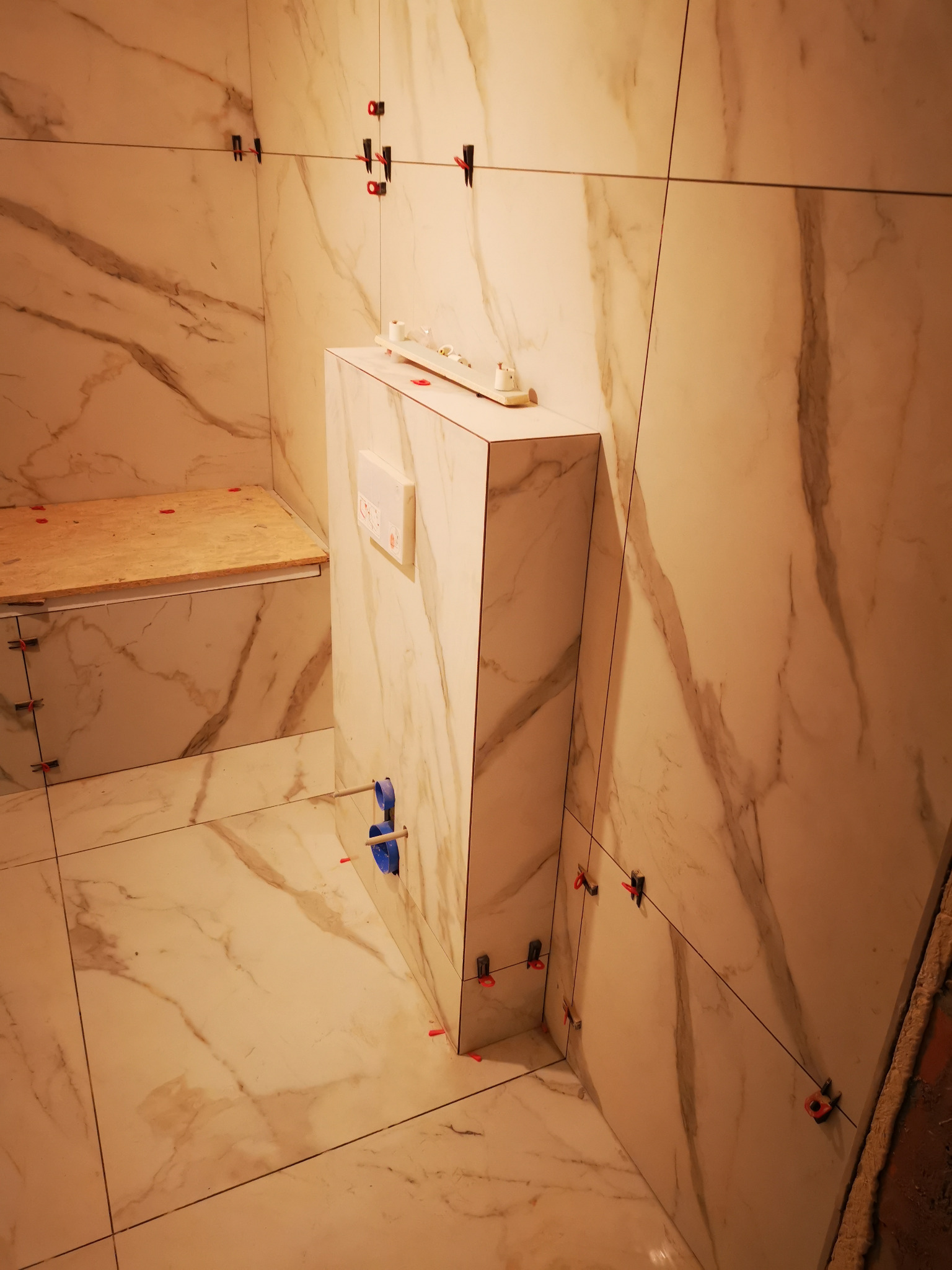 Świeżo ułożone, marmurowe płytki w łazience podczas prac wykończeniowych; widoczne elementy montażowe, stelaż podtynkowy i fragment konstrukcji pod półkę.