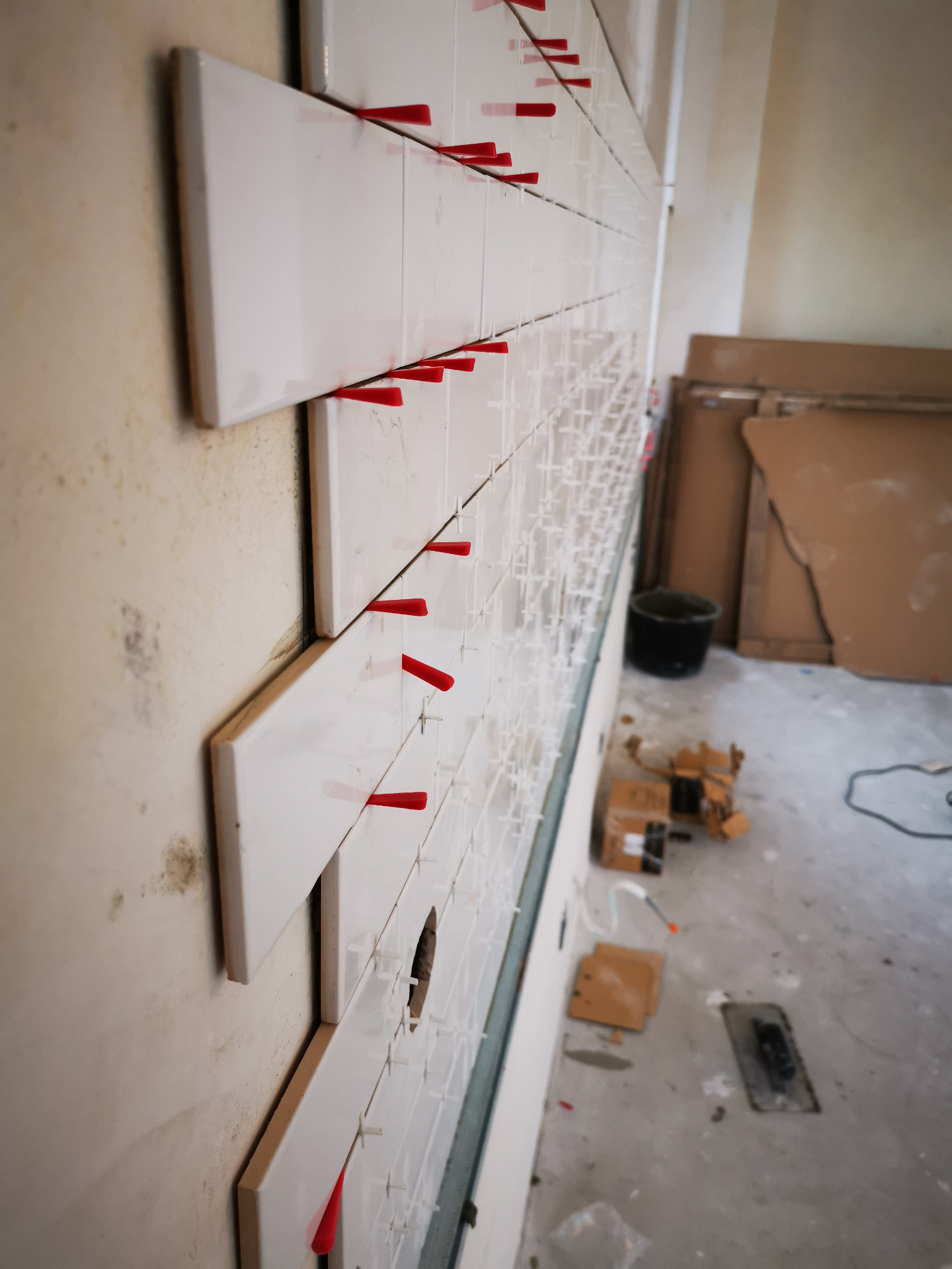 Układanie białych, prostokątnych płytek na ścianie z użyciem czerwonych klinów dystansowych, widoczny otwór w ścianie, narzędzia glazurnicze i kartony na podłodze.