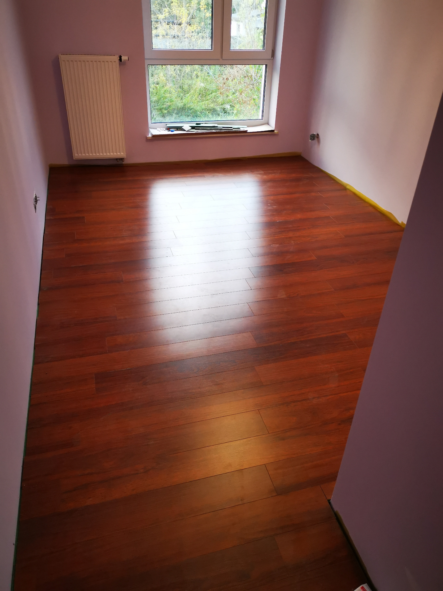 Pokój po remoncie z nową, błyszczącą podłogą z paneli imitujących drewno, ściany pomalowane na jasny fiolet, zabezpieczone taśmą malarską przy podłodze, biały grzejnik pod oknem.