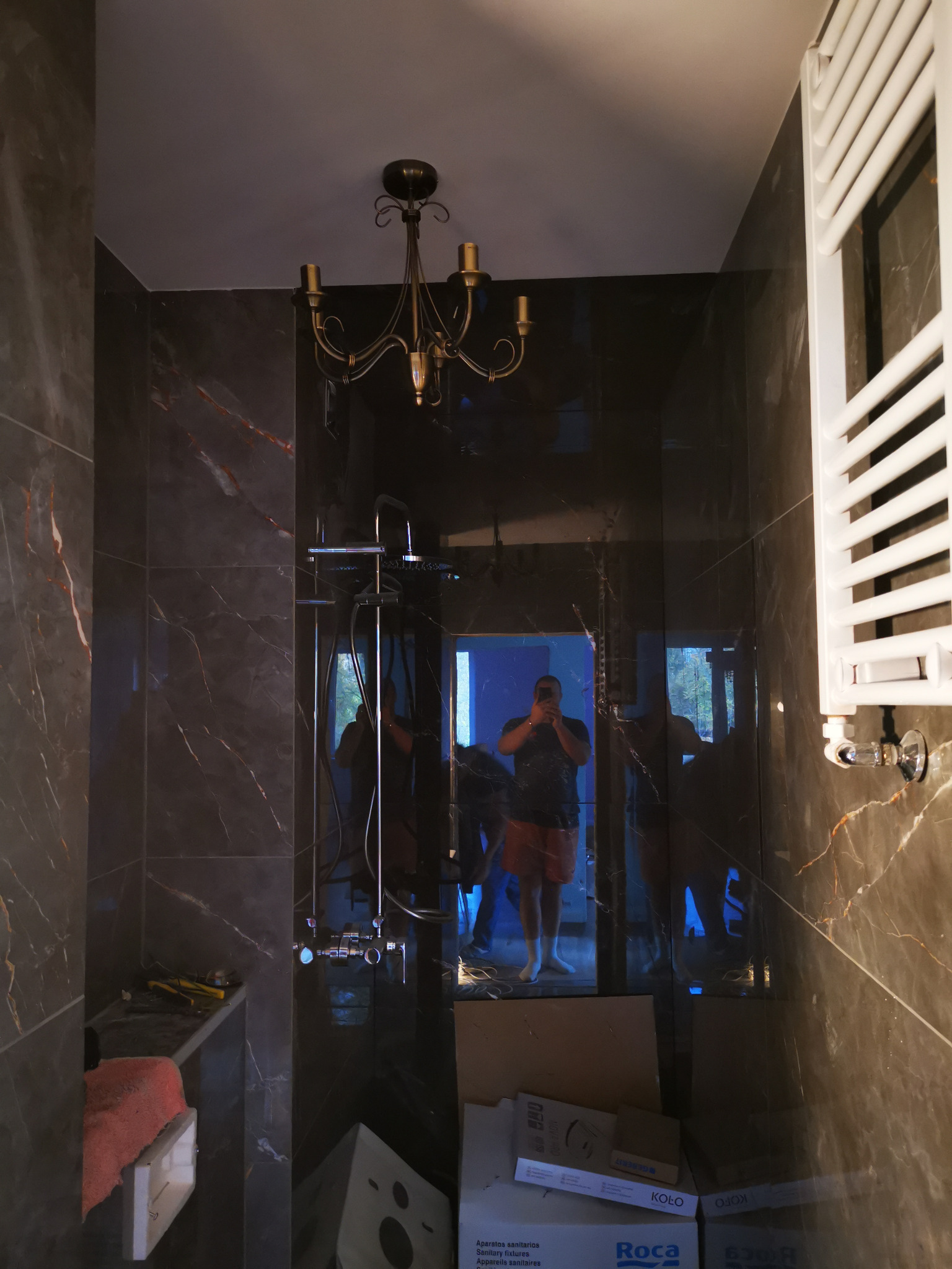 Wykończona łazienka z ciemnymi płytkami imitującymi marmur, widoczna bateria prysznicowa, grzejnik drabinkowy, ozdobny żyrandol i odbicie osoby robiącej zdjęcie. W tle widoczne kartony i narzędzia.