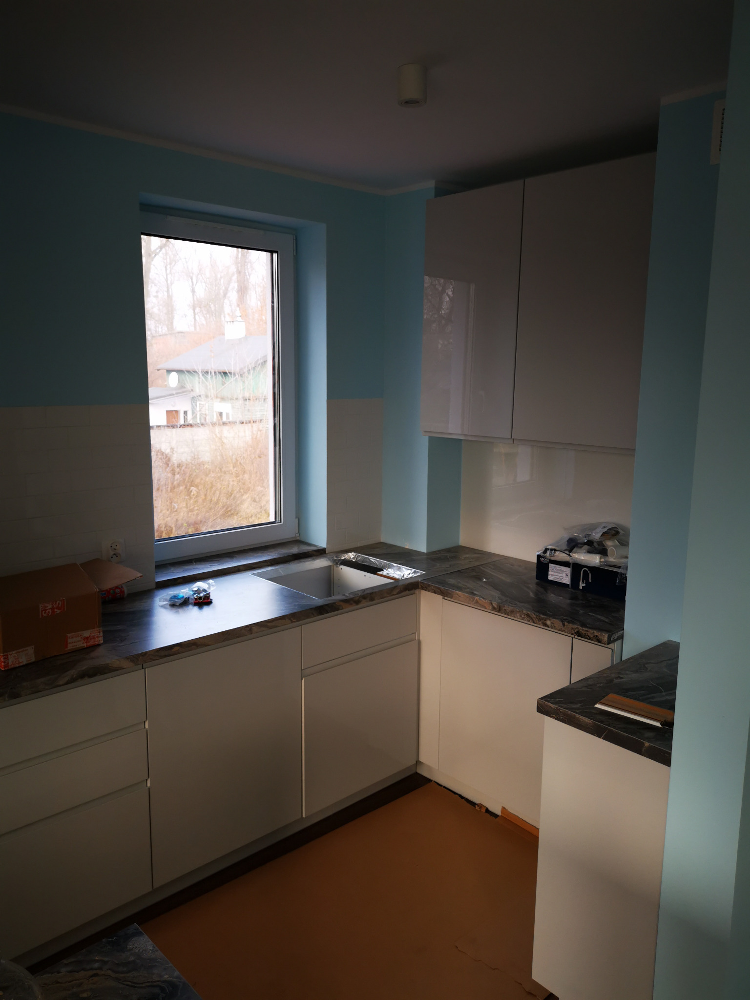 Nowoczesna kuchnia z białymi szafkami i blatem imitującym marmur, okno z widokiem na dom, w tle błękitne ściany, na blacie kartonowe pudełko i akcesoria do montażu zlewu.