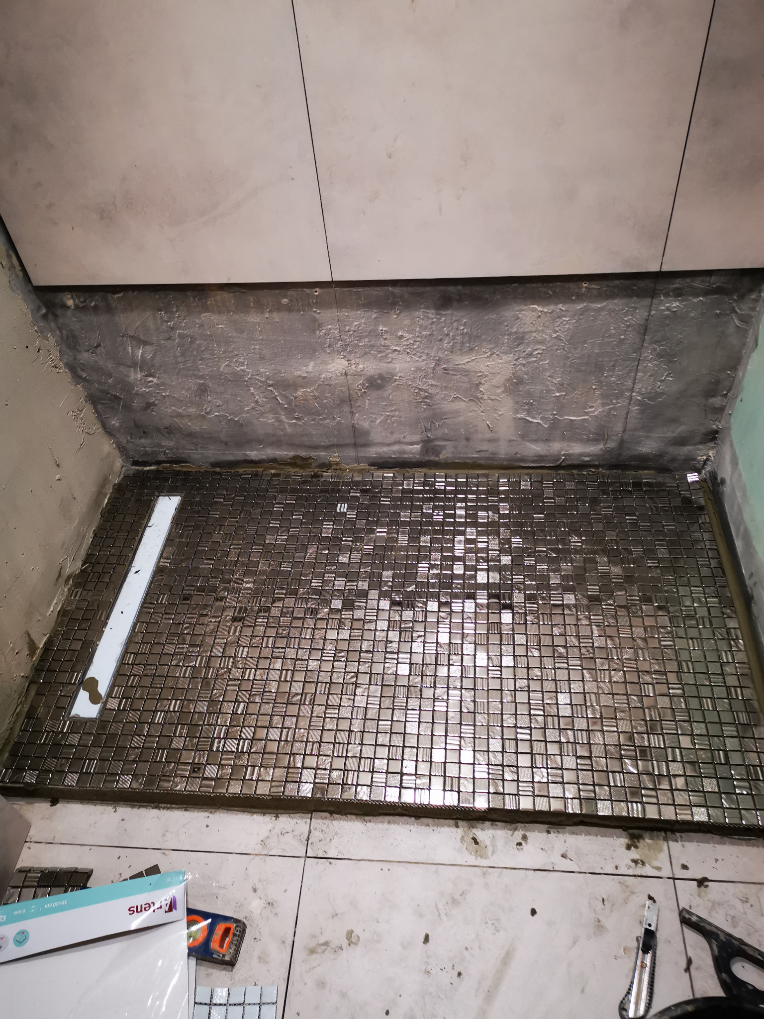 Przygotowanie do wyłożenia brodzika mozaiką w łazience, widoczne narzędzia, poziomica, opakowanie płytek i nożyk introligatorski.