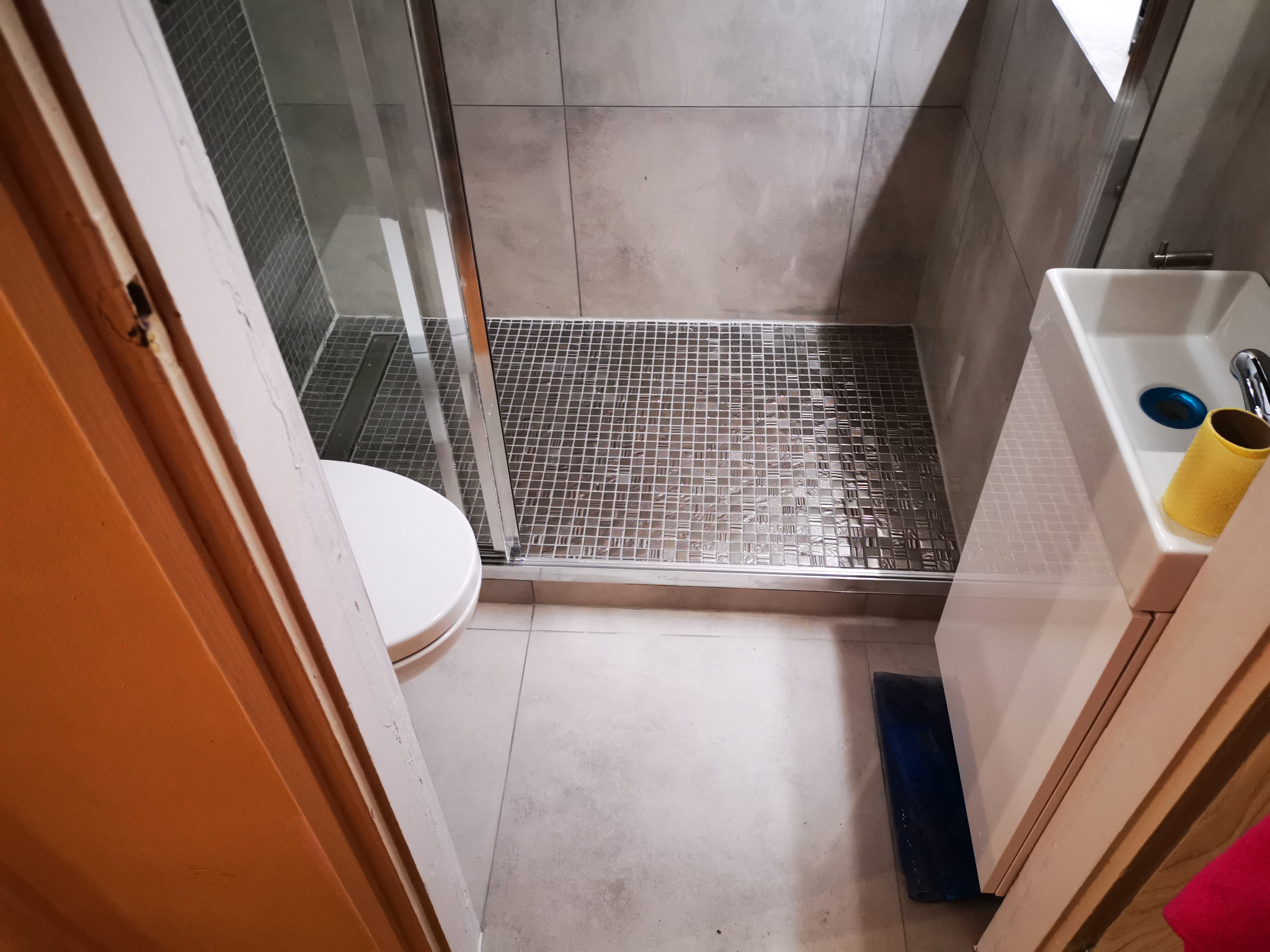 Widok z góry na wyremontowaną łazienkę z kabiną prysznicową wyłożoną mozaiką, umywalką z żółtym pojemnikiem i toaletą, widzianą przez otwarte drzwi.