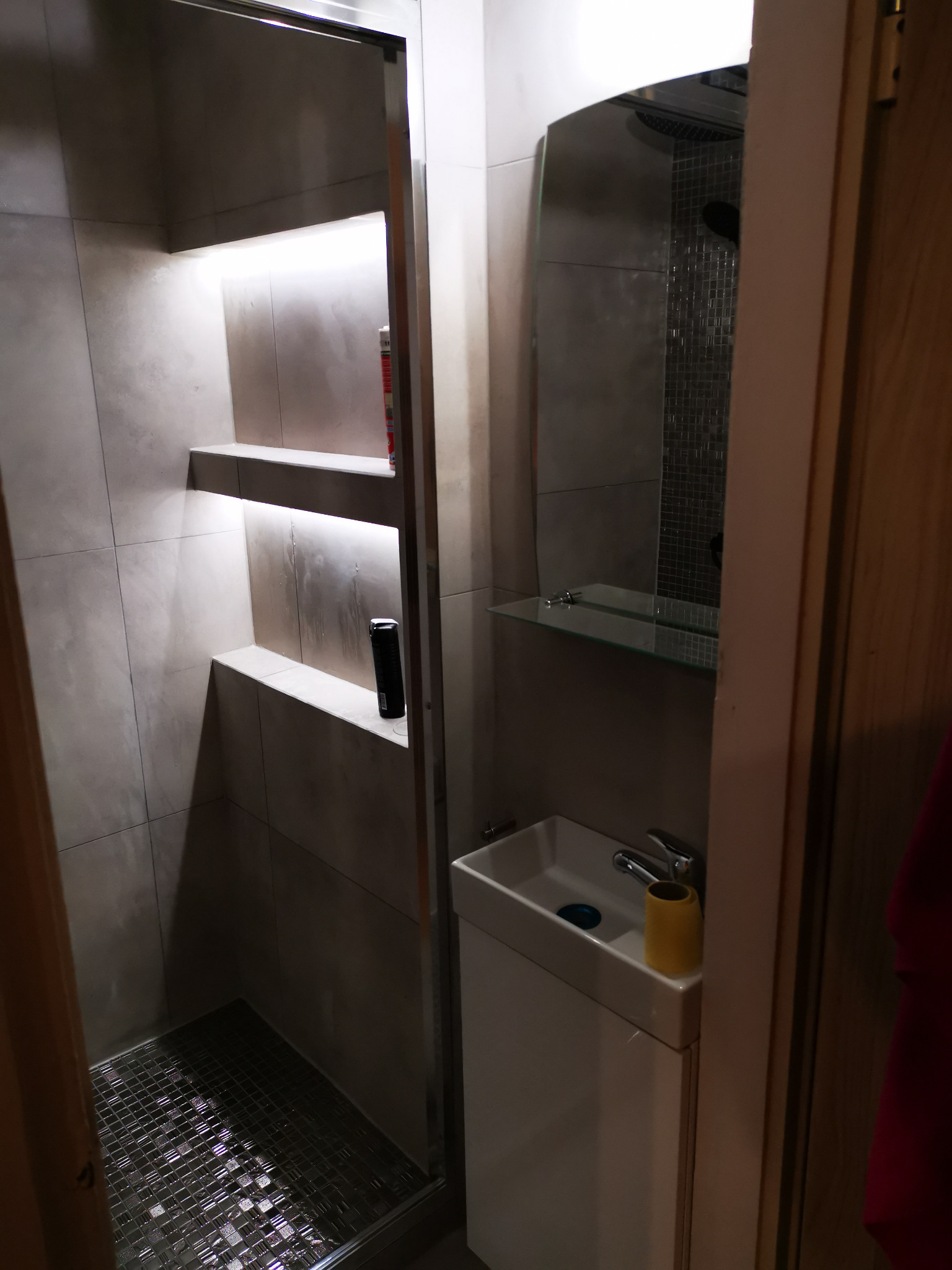 Nowoczesna łazienka po remoncie: kabina prysznicowa z mozaikową podłogą, wnęki z podświetleniem i kosmetykami, umywalka nablatowa z żółtym pojemnikiem na szczoteczki.