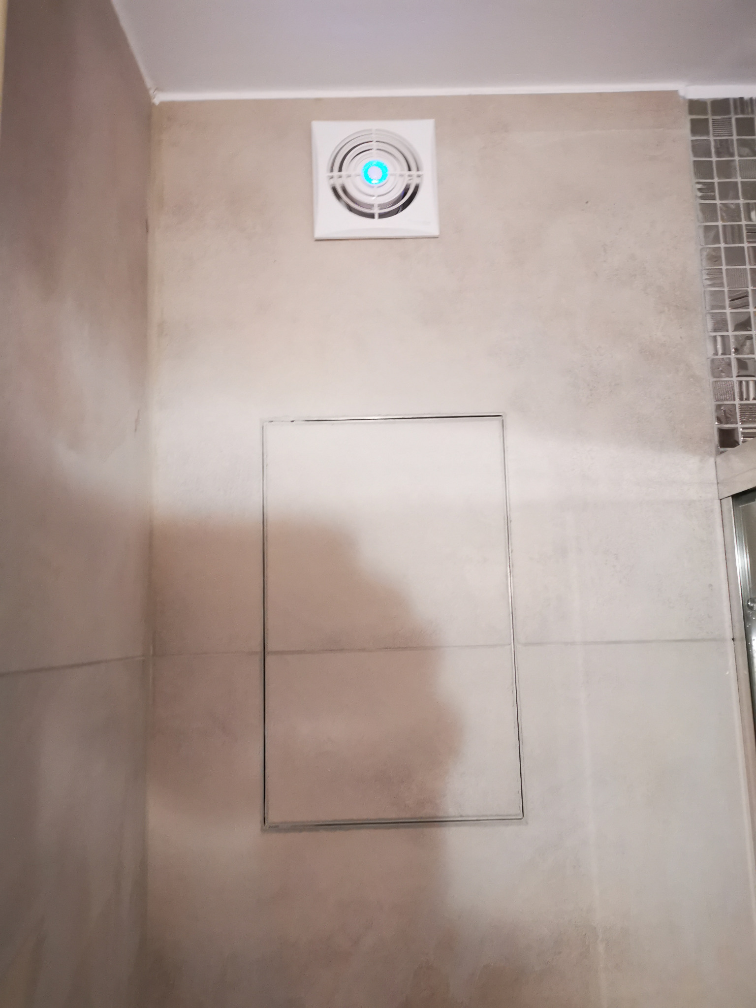 Wykończona ściana łazienki z beżowych płytek z ukrytym włazem rewizyjnym i wentylatorem sufitowym z niebieskim podświetleniem LED.