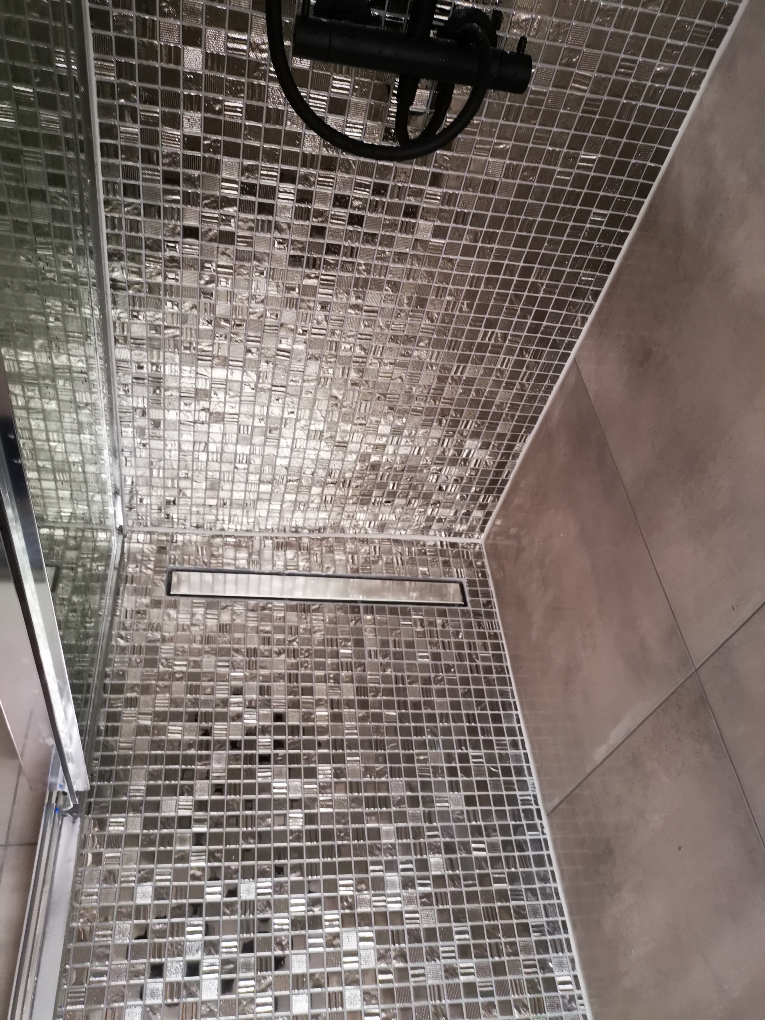 Kabina prysznicowa wyłożona mozaiką w odcieniach szarości i srebra, z odpływem liniowym w podłodze i deszczownicą zamontowaną na suficie.