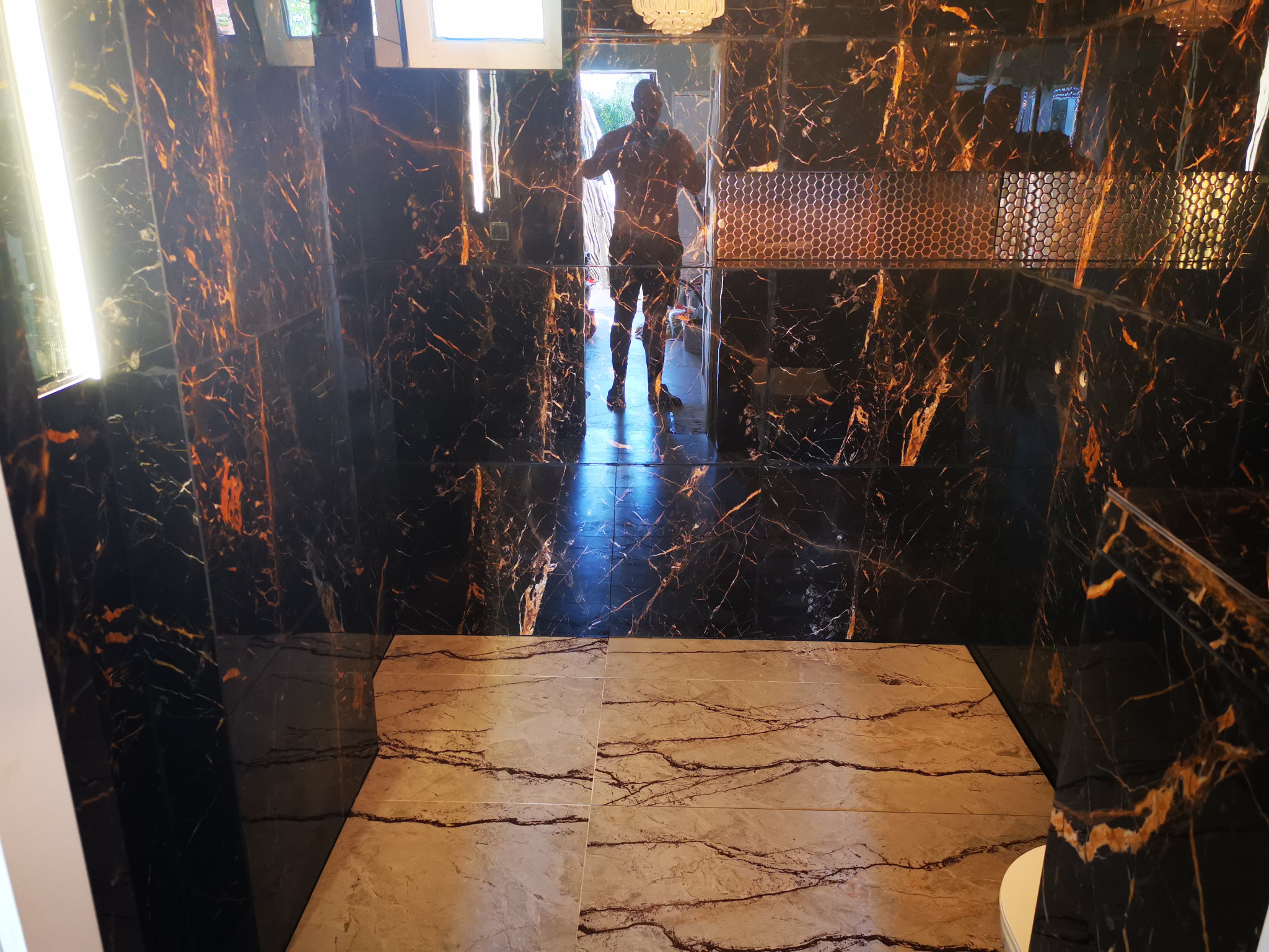 Luksusowa łazienka wykończona czarnym marmurem z złotymi żyłami na ścianach i beżowymi płytkami podłogowymi, z widocznym odbiciem fotografa w lustrzanej powierzchni płytek.