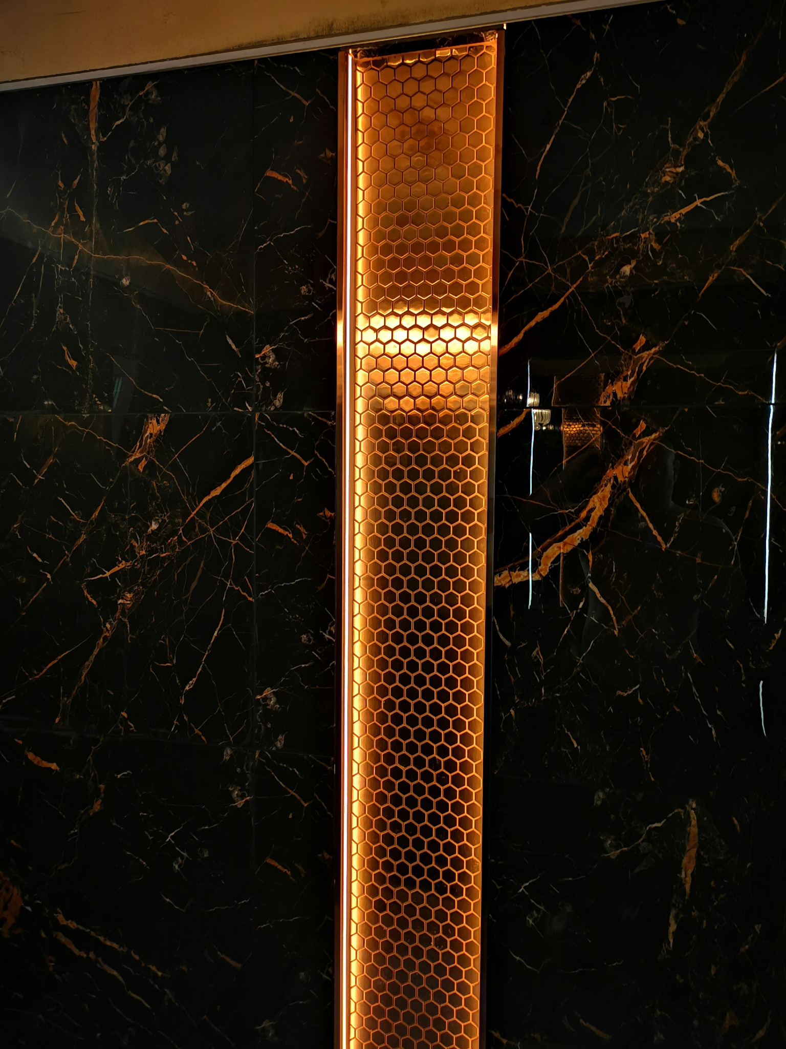 Ściana wykończona czarnym marmurem z żyłami w kolorze miedzi, obok pionowy panel dekoracyjny z podświetlaną strukturą plastra miodu.