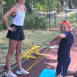 MKrawczyńska team - Kobieta w sportowym stroju nadzoruje chłopca ćwiczącego z gumą na niebieskiej macie, w tle żółte płotki treningowe.