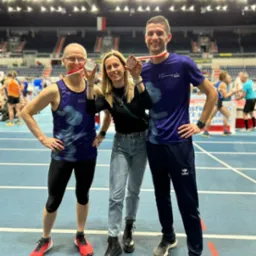 Zdjęcie z Gdańska przedstawia trenera biegania i dwóch biegaczy z medalami na szyjach, stojących na bieżni w hali sportowej, po zakończonym biegu.