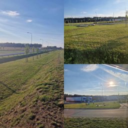 FLY PARADISE S.C - Widok na pas zieleni wzdłuż autostrady z młodymi drzewkami, fragment parkingu dla tirów w oddali, błękitne niebo z chmurami.