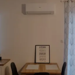 Biały klimatyzator Haier zamontowany nad drewnianym stołem jadalnym z trzema krzesłami, dekoracyjna zasłona w tle.