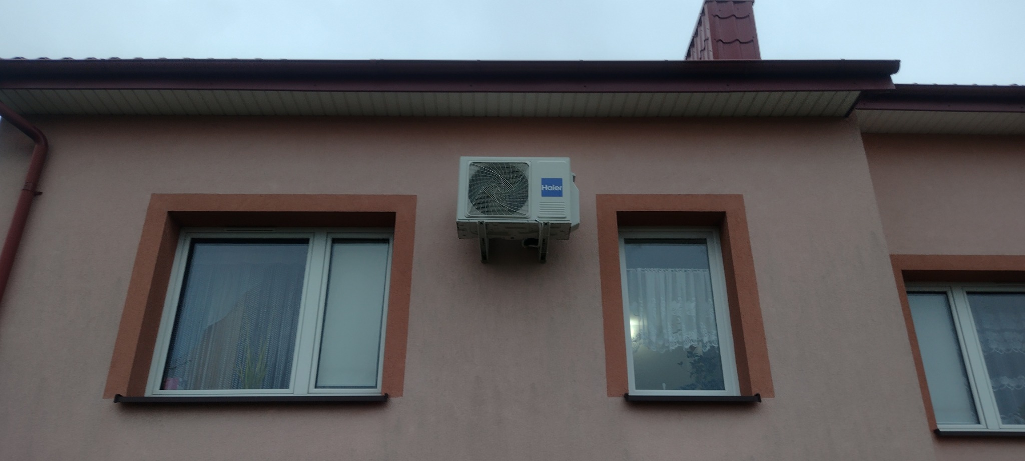 Biała jednostka zewnętrzna klimatyzatora Haier zamontowana na elewacji domu w kolorze pudrowego różu, widoczna pomiędzy dwoma oknami z brązowymi ramami i białymi parapetami, na tle pochmurnego nieba.