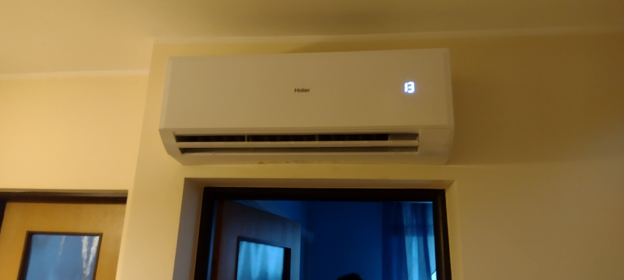 Biały klimatyzator Haier zamontowany na ścianie nad drzwiami, wyświetlający temperaturę 13 stopni.
