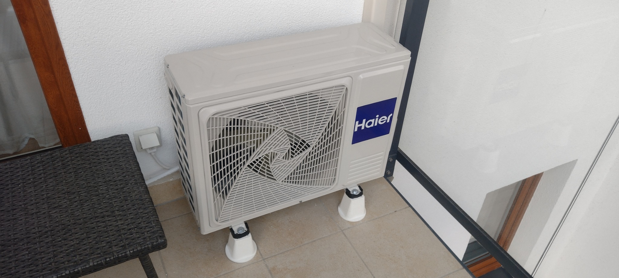 Zewnętrzna jednostka klimatyzacji Haier zamontowana na balkonie, widoczne białe wsporniki i kratka wentylacyjna z geometrycznym wzorem.
