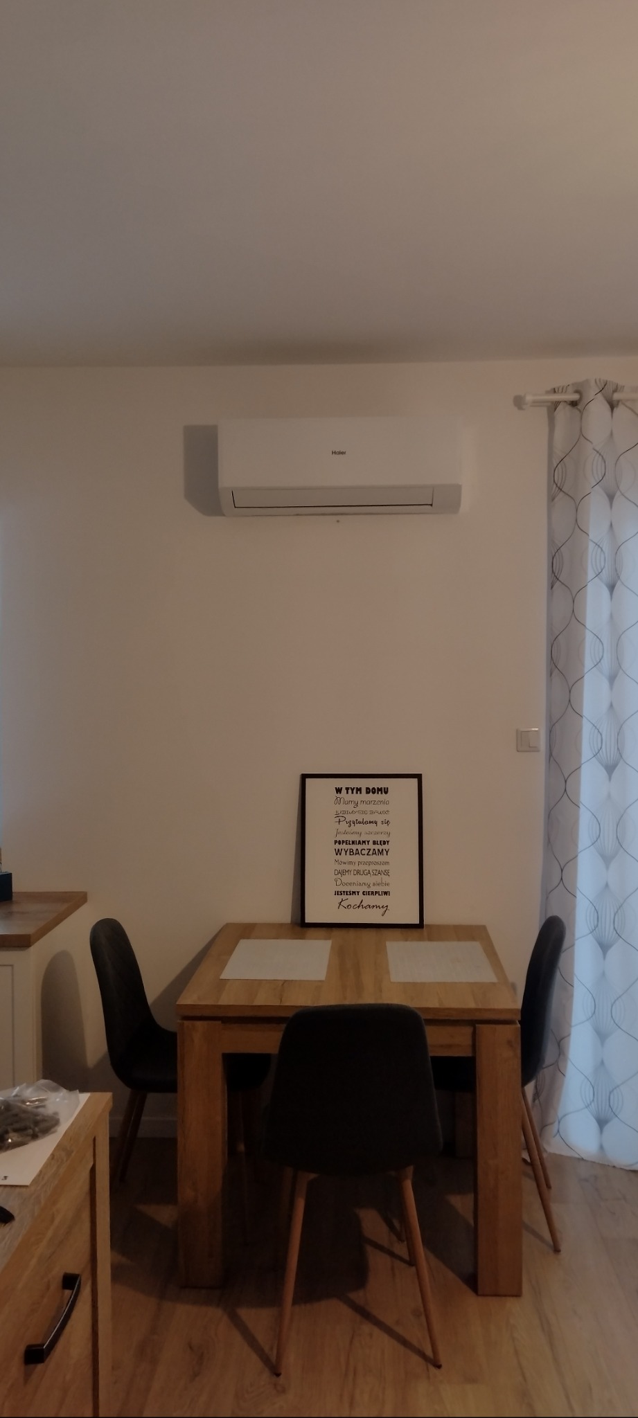 Biały klimatyzator Haier zamontowany nad drewnianym stołem jadalnym z trzema krzesłami, dekoracyjna zasłona w tle.