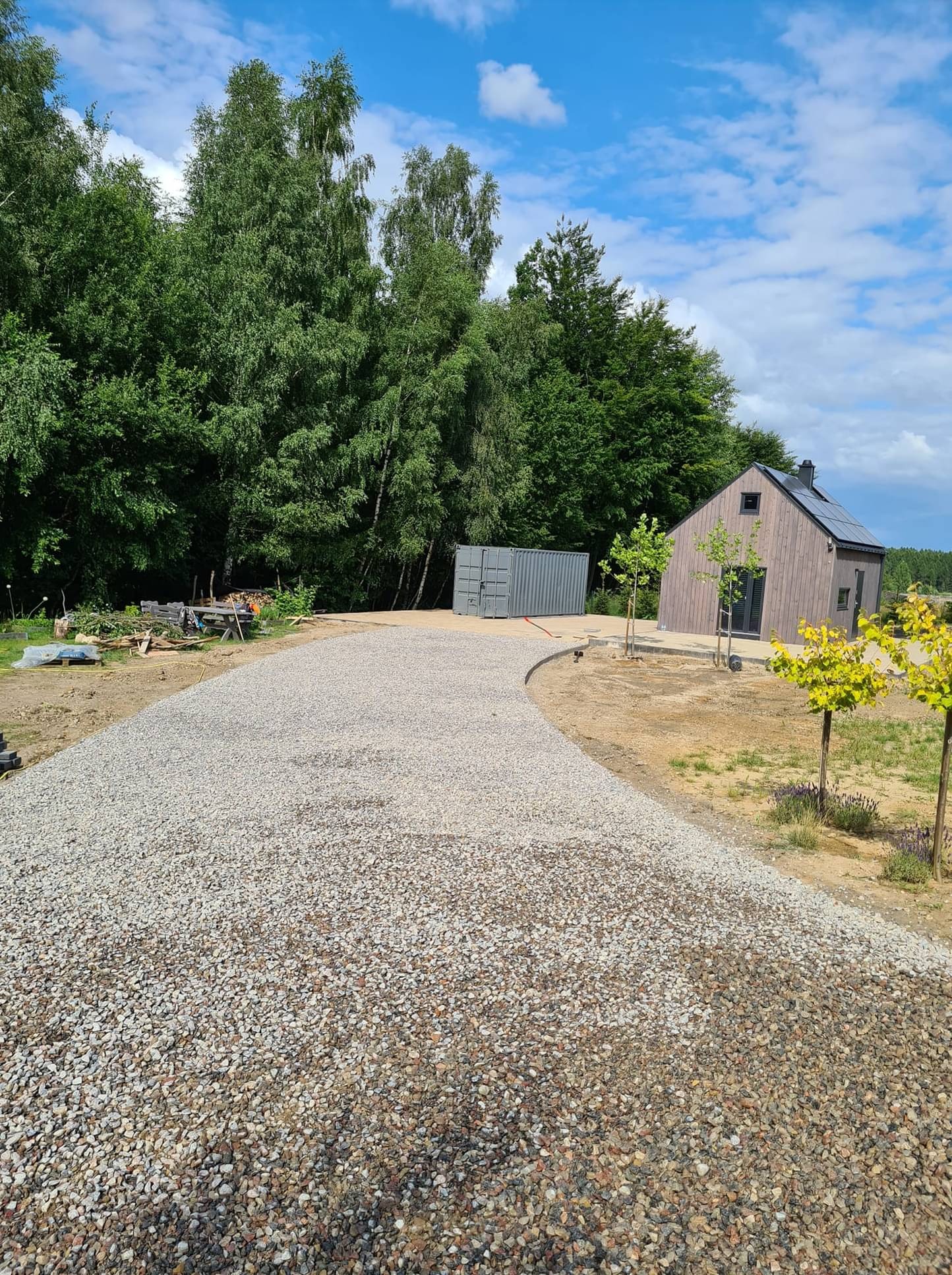 Nowa, żwirowa droga do nowoczesnego domu z panelami słonecznymi, z kontenerem i przygotowanym miejscem na drewno w tle, otoczona zielenią.