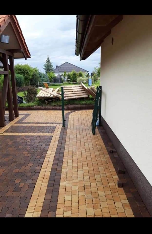 Ukończona nawierzchnia z dwukolorowej kostki brukowej ułożonej w pasy, z widocznymi elementami krajobrazu w tle i stertą desek.